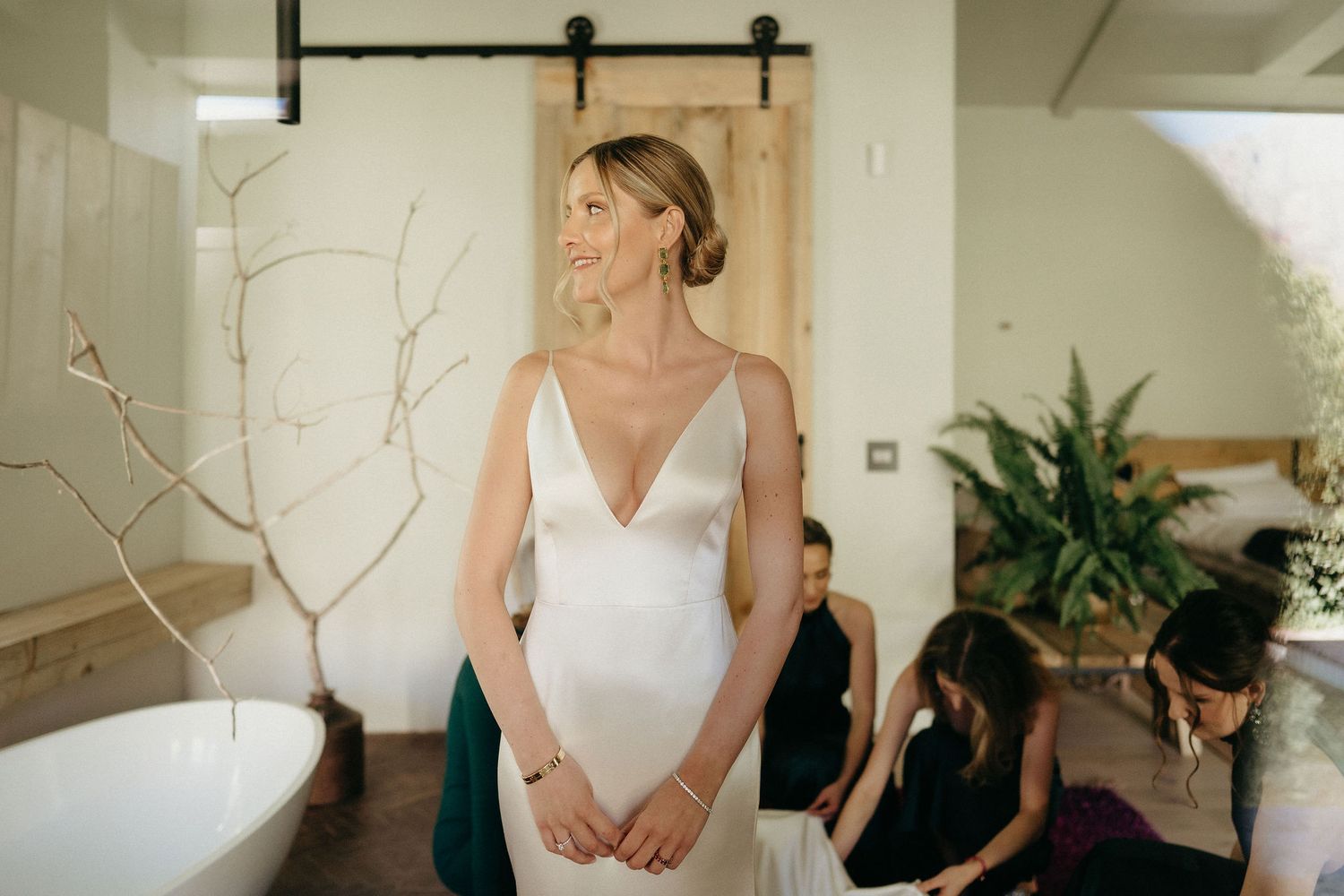 A person in an elegant white wedding dress stands in a bathroom with a sliding barn door and potted plant.