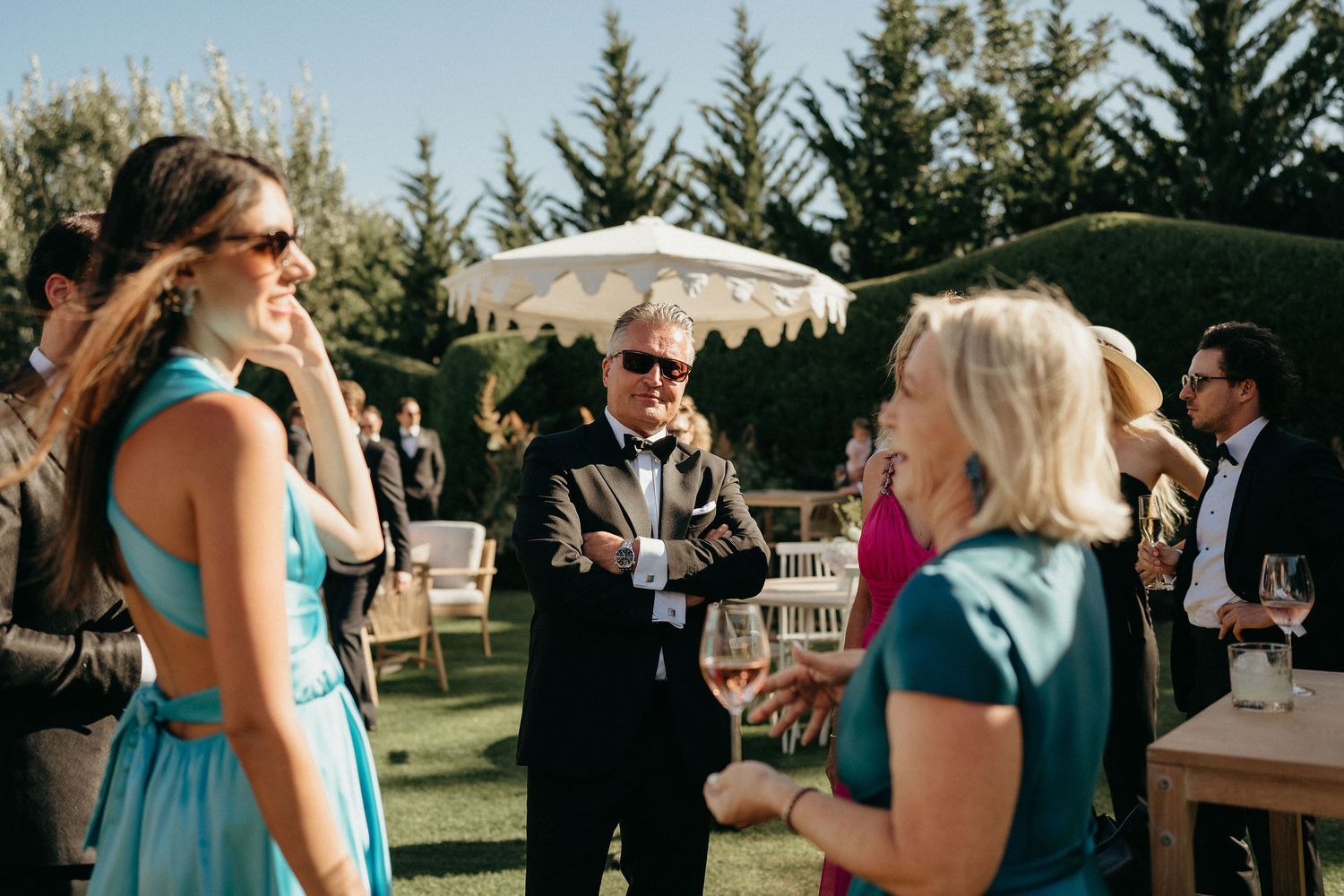 A wedding reception celebration with guests mingling outdoors under white umbrellas and trees.