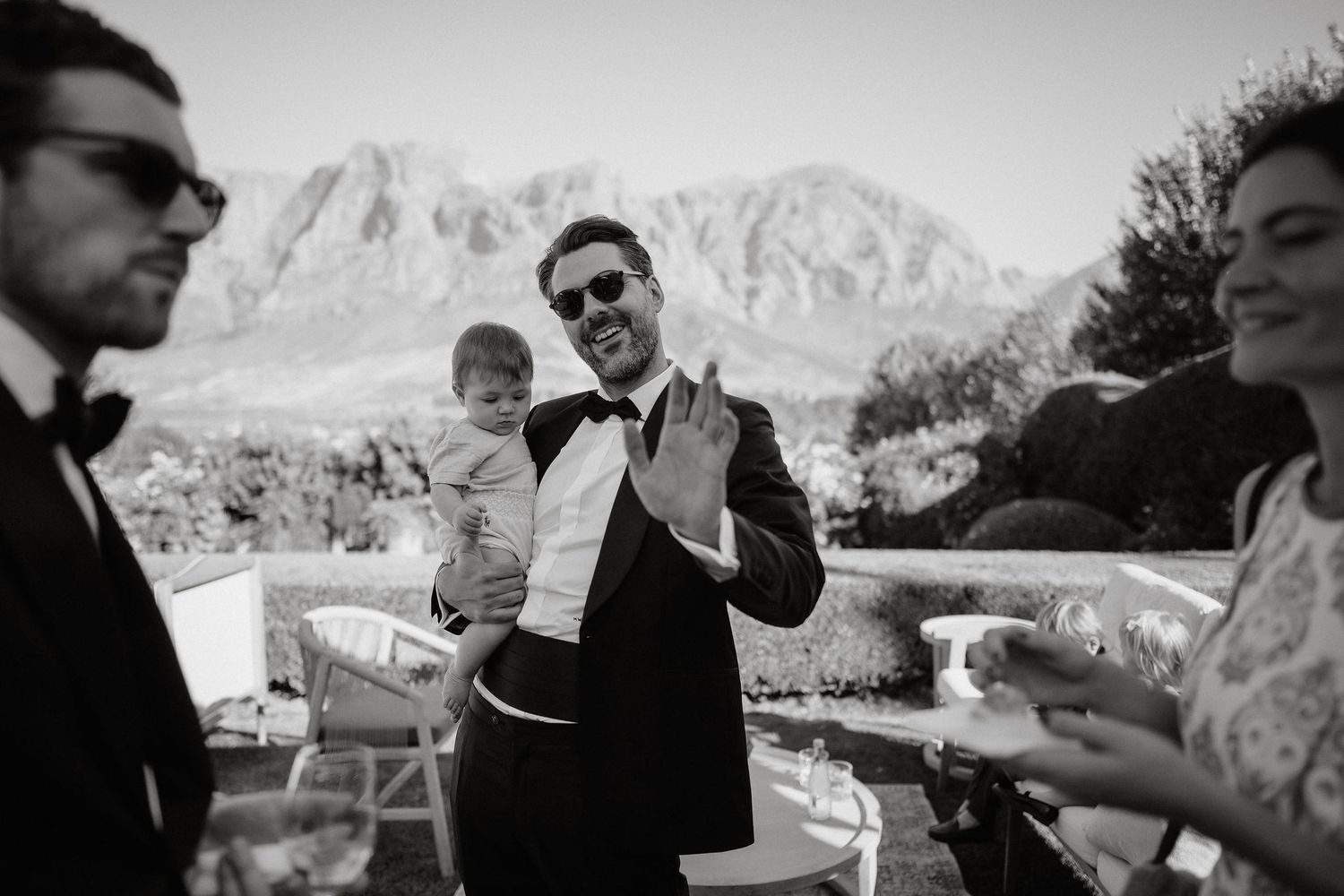 A black and white outdoor gathering with people in formal attire against a scenic mountain backdrop.