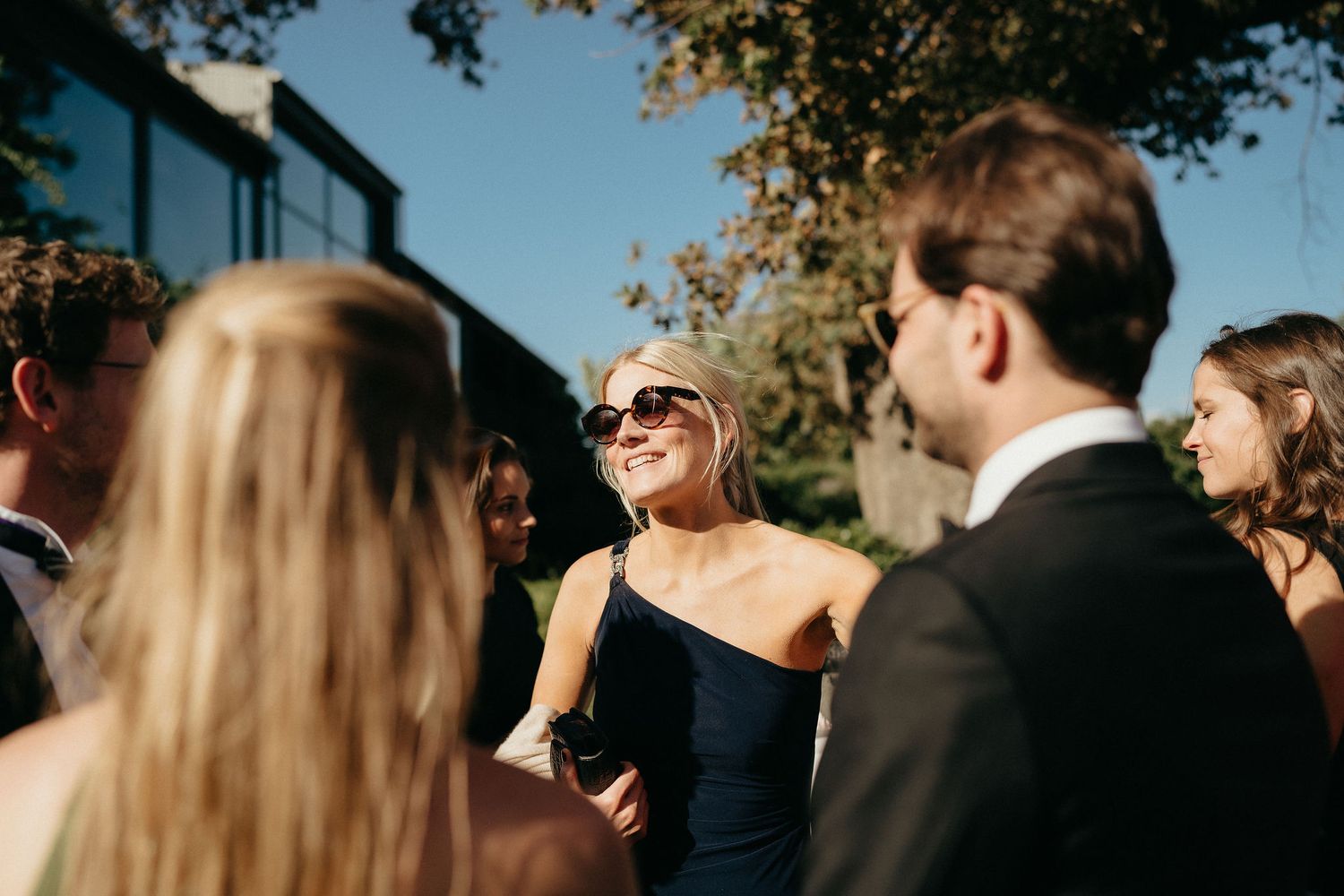 A candid moment at an outdoor social gathering shows people laughing and conversing in warm sunlight.