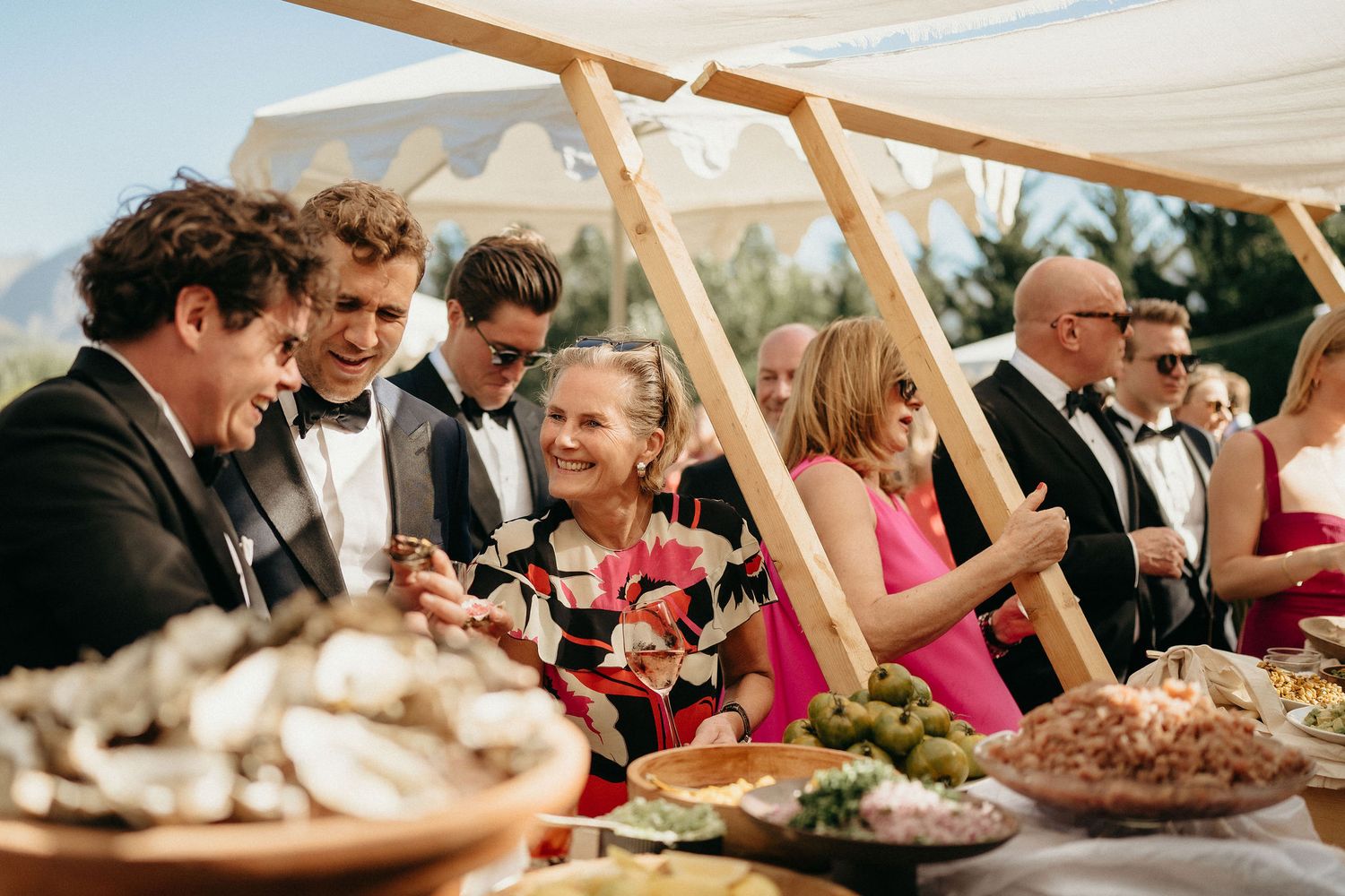 Wedding guests seated at long tables under a white tent enjoy a festive outdoor reception meal together.