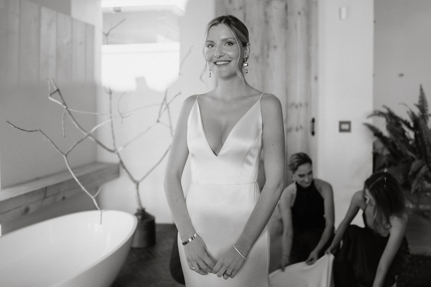 A bride in an elegant white gown smiles brightly in a bright indoor setting with natural light streaming through windows.