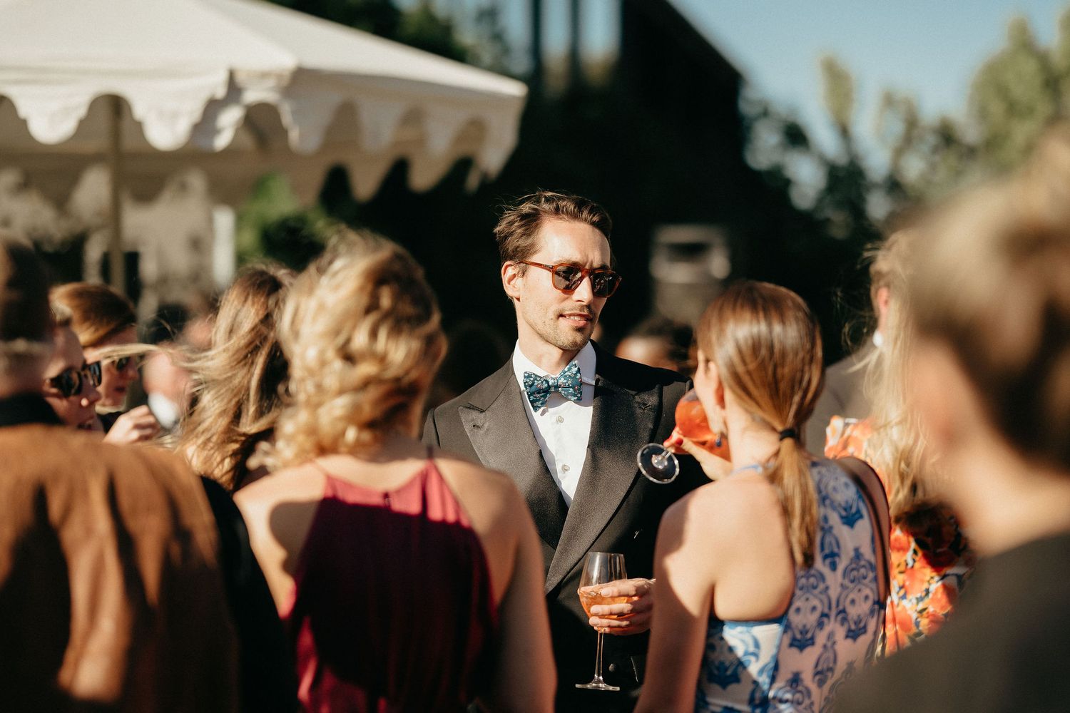 Wedding guests enjoy cocktail hour on an outdoor patio with string lights overhead during sunset.