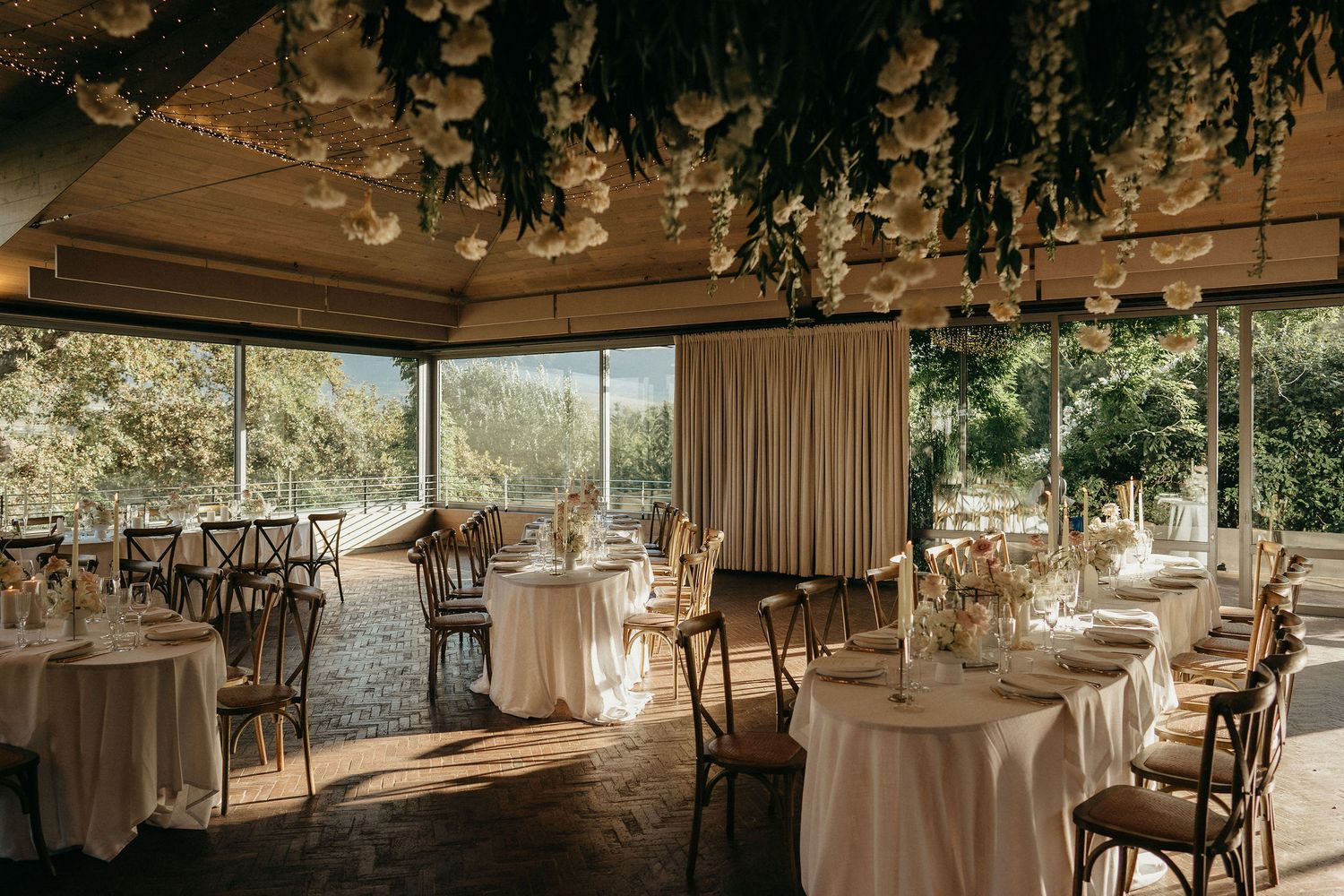 Elegant wedding reception venue decorated with hanging greenery, white linens, and wooden chairs in a rustic indoor setting.