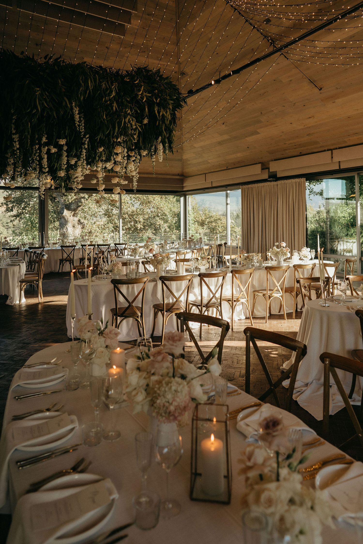 Elegant rustic wedding reception venue with wooden cross-back chairs and white table settings in a warm-lit indoor space.