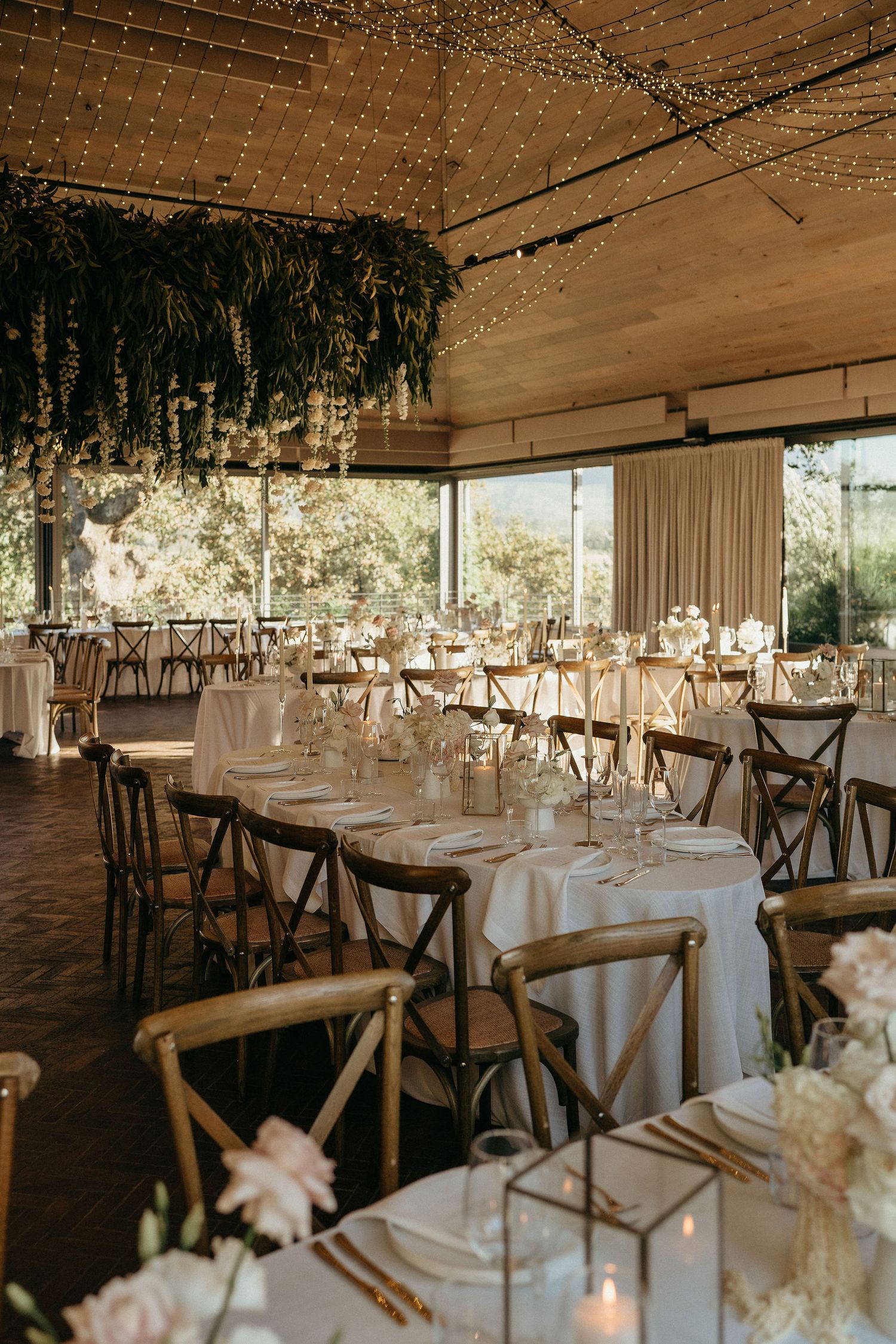 Elegant wedding reception venue with white tablecloths, wooden cross-back chairs, and twinkling lights under a rustic ceiling.