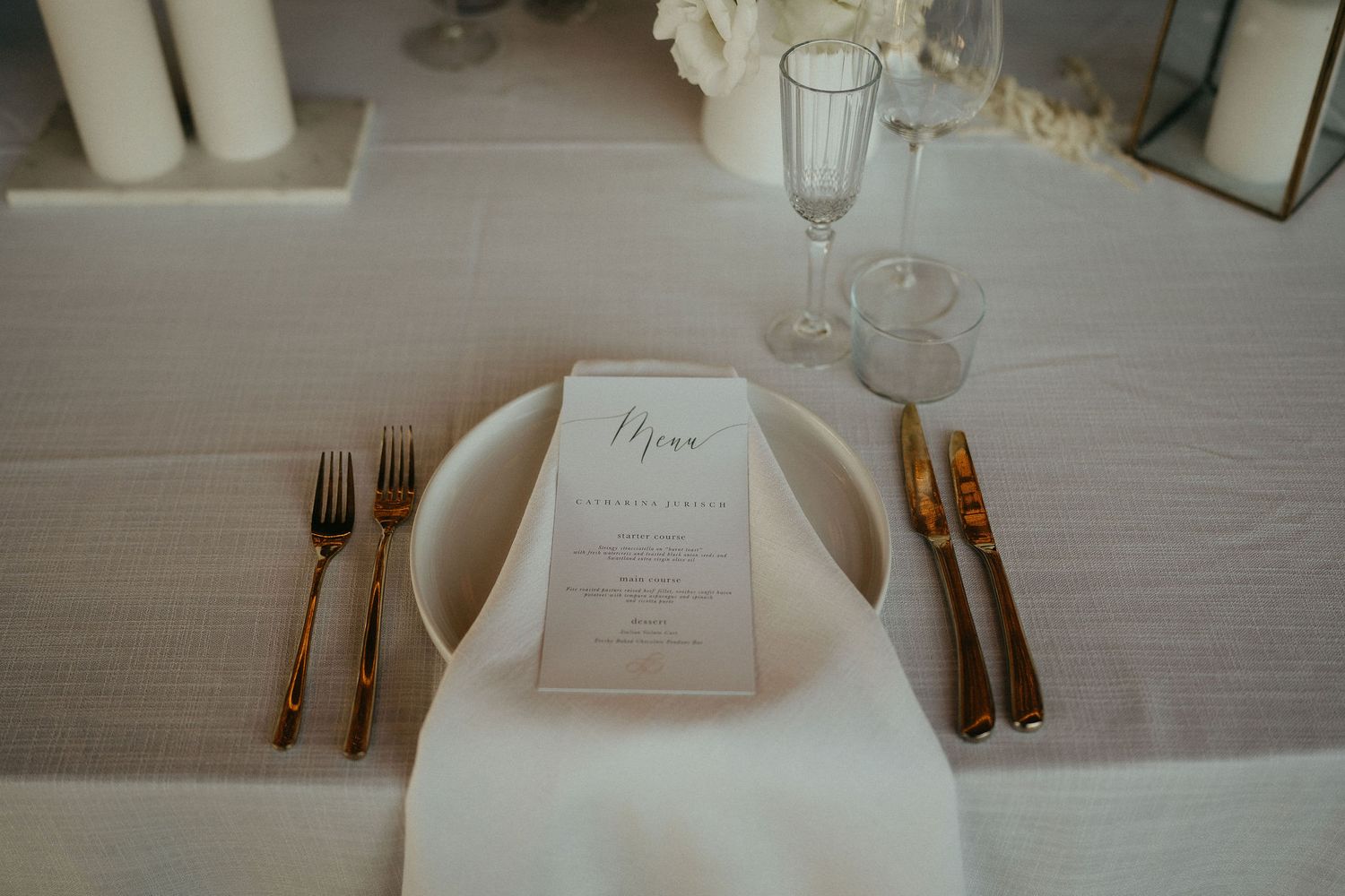Elegant place setting with menu card, white plate, and gold cutlery on crisp white tablecloth.
