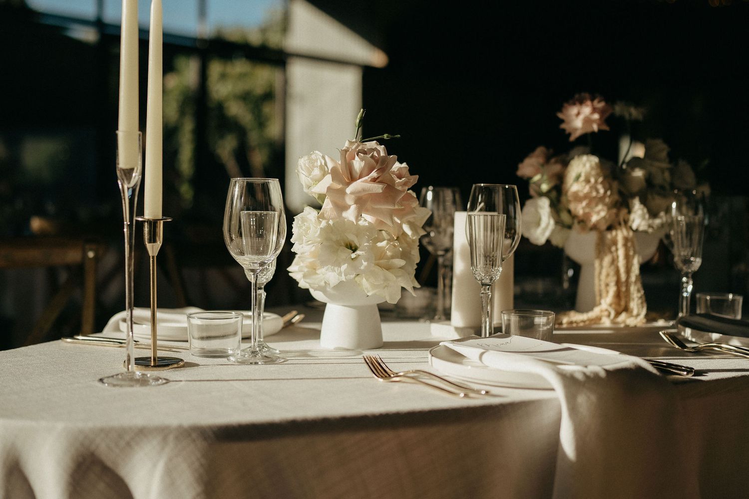 Romantic wedding table setting with tall candles, wine glasses and floral centerpiece in evening light.