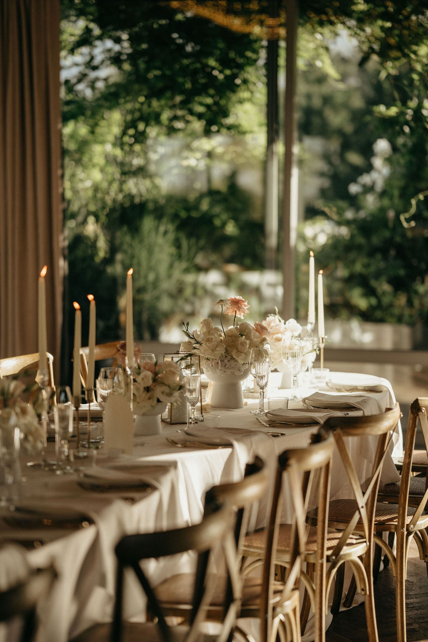 Elegant wedding reception tables set with white linens, candles and rustic wooden chairs in a natural light-filled venue.