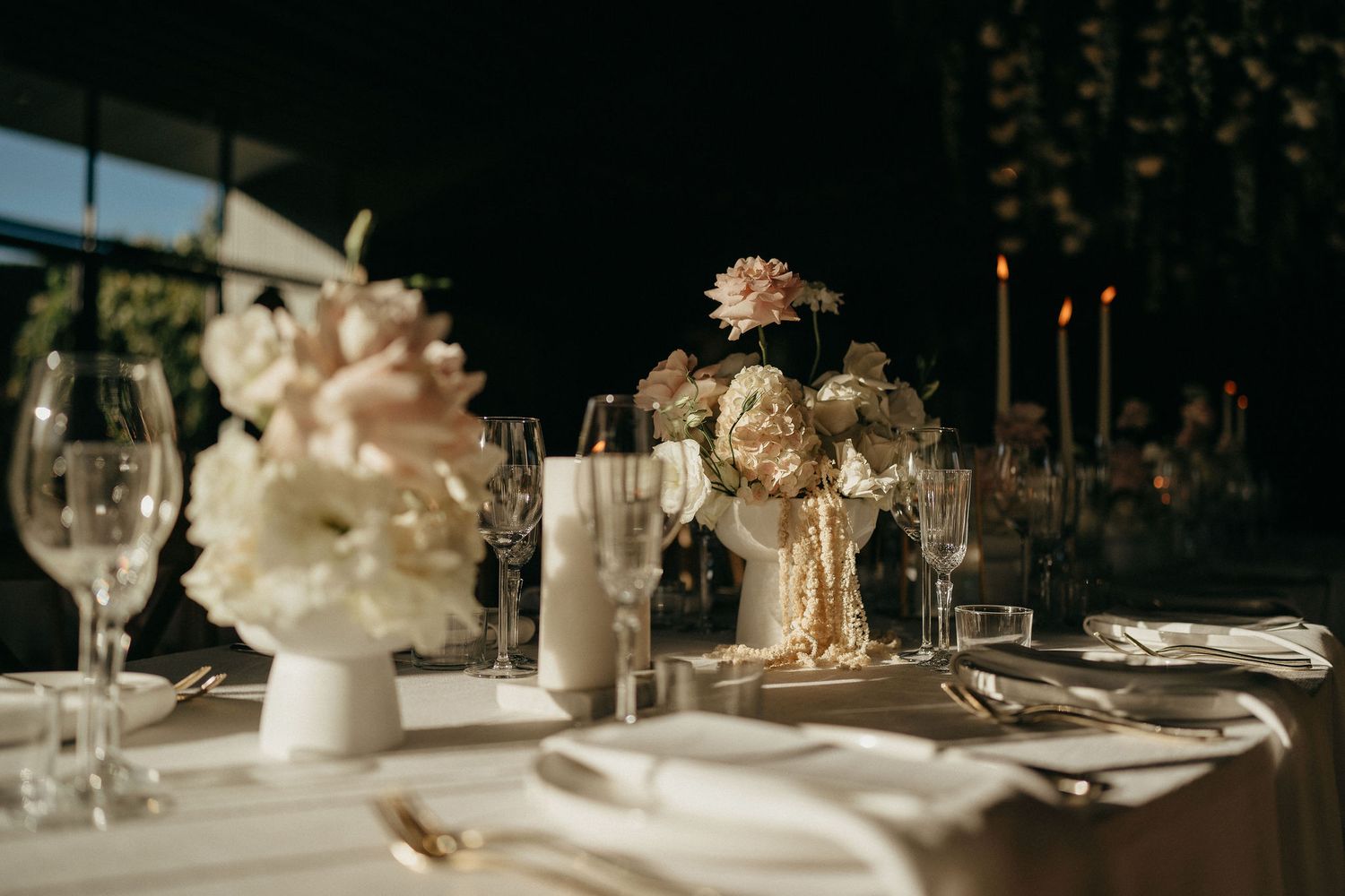Elegant wedding table setting with white floral centerpieces, candlesticks and crystal glassware in warm lighting.