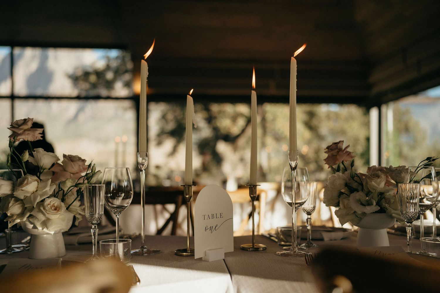 Elegant wedding table setting with tall taper candles and floral centerpieces in warm sunset lighting.