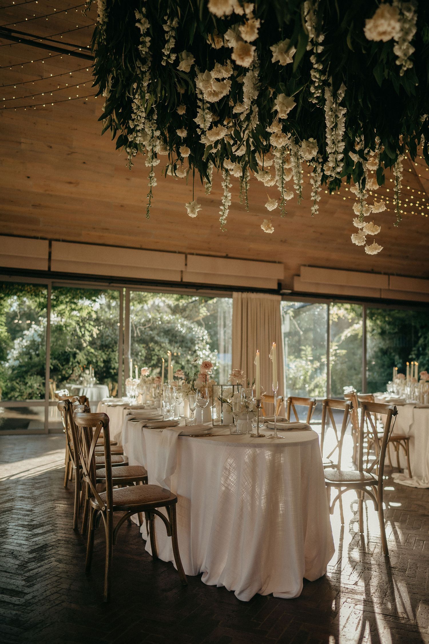 Elegant wedding reception venue with white tablecloths, wooden chairs, and hanging floral arrangements under rustic wooden beams.