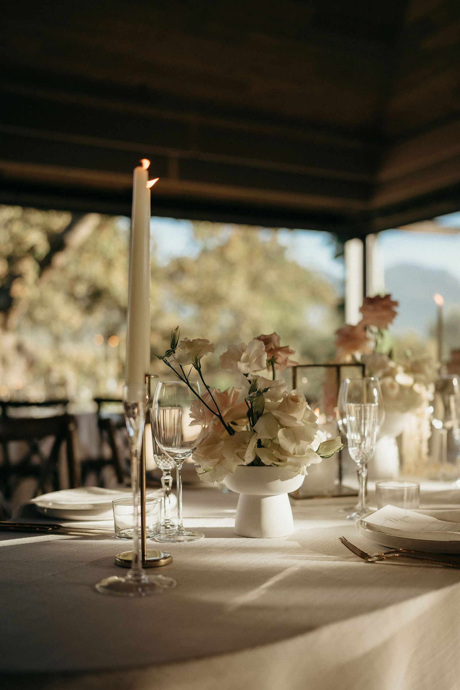 Elegant wedding reception table settings with white floral centerpieces and tall candles in a sunlit venue.
