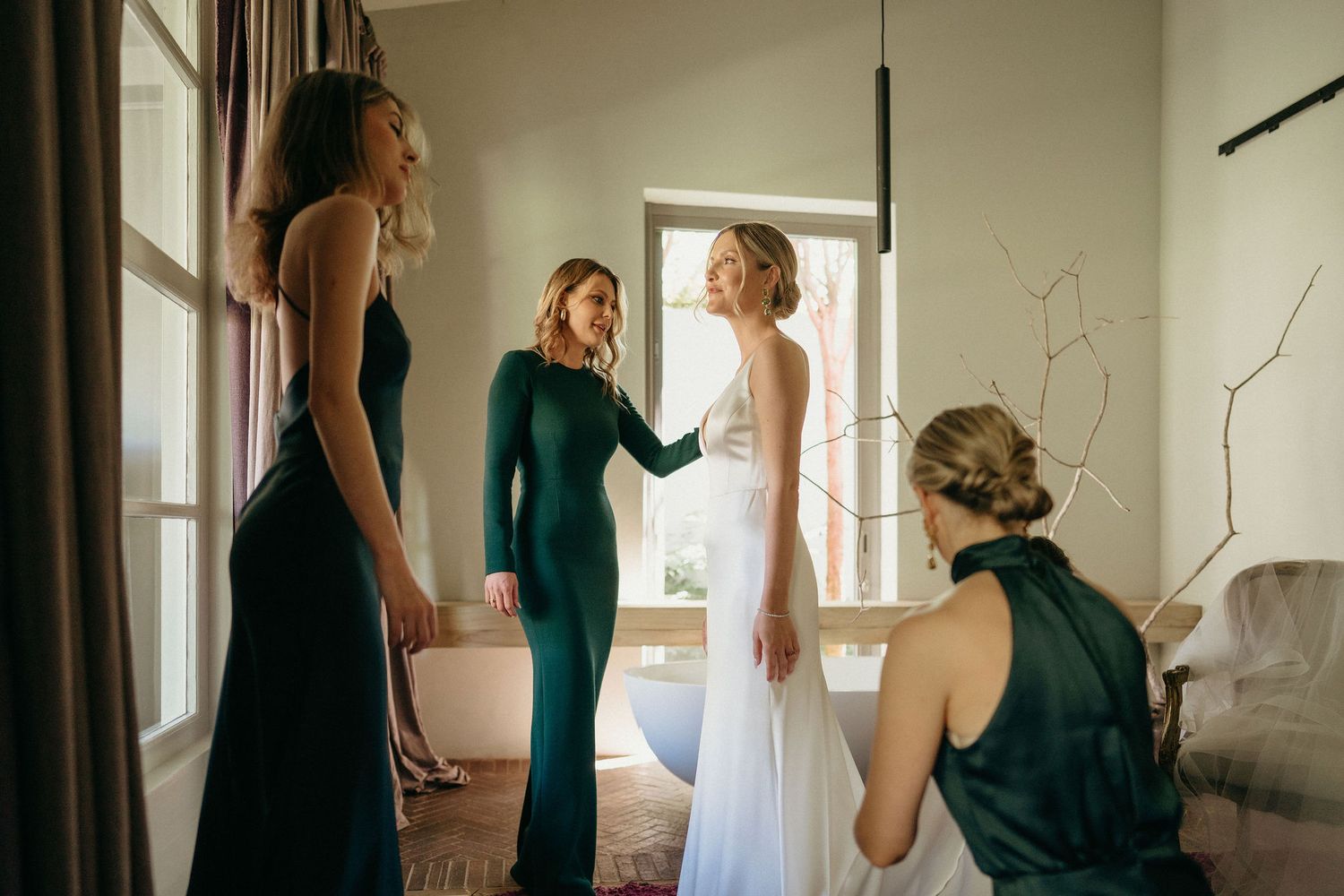 A bride in a white gown gets ready with her bridesmaids wearing elegant dark dresses in a sunlit room.