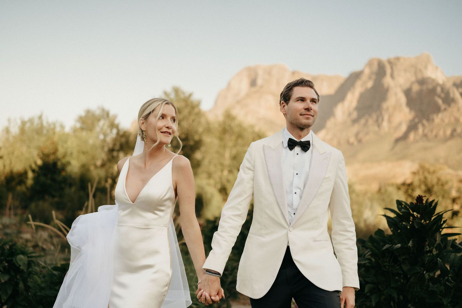 A couple in wedding attire stands against a backdrop of majestic mountain peaks during a golden sunset.