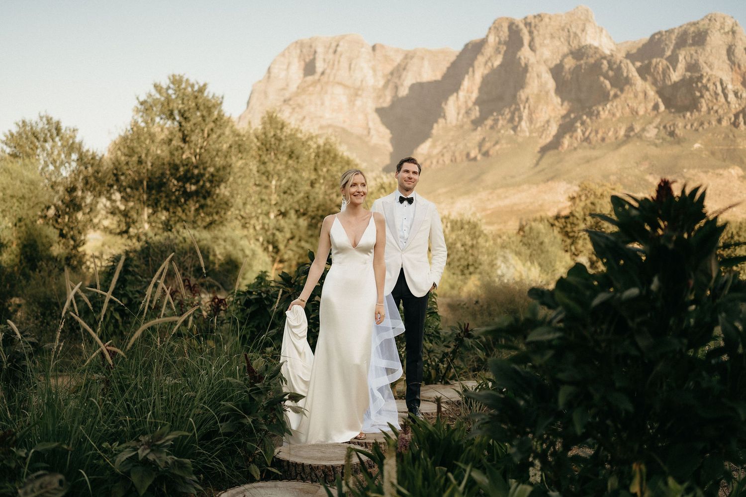 A couple in wedding attire stands together against a scenic mountain backdrop in the desert landscape at sunset.