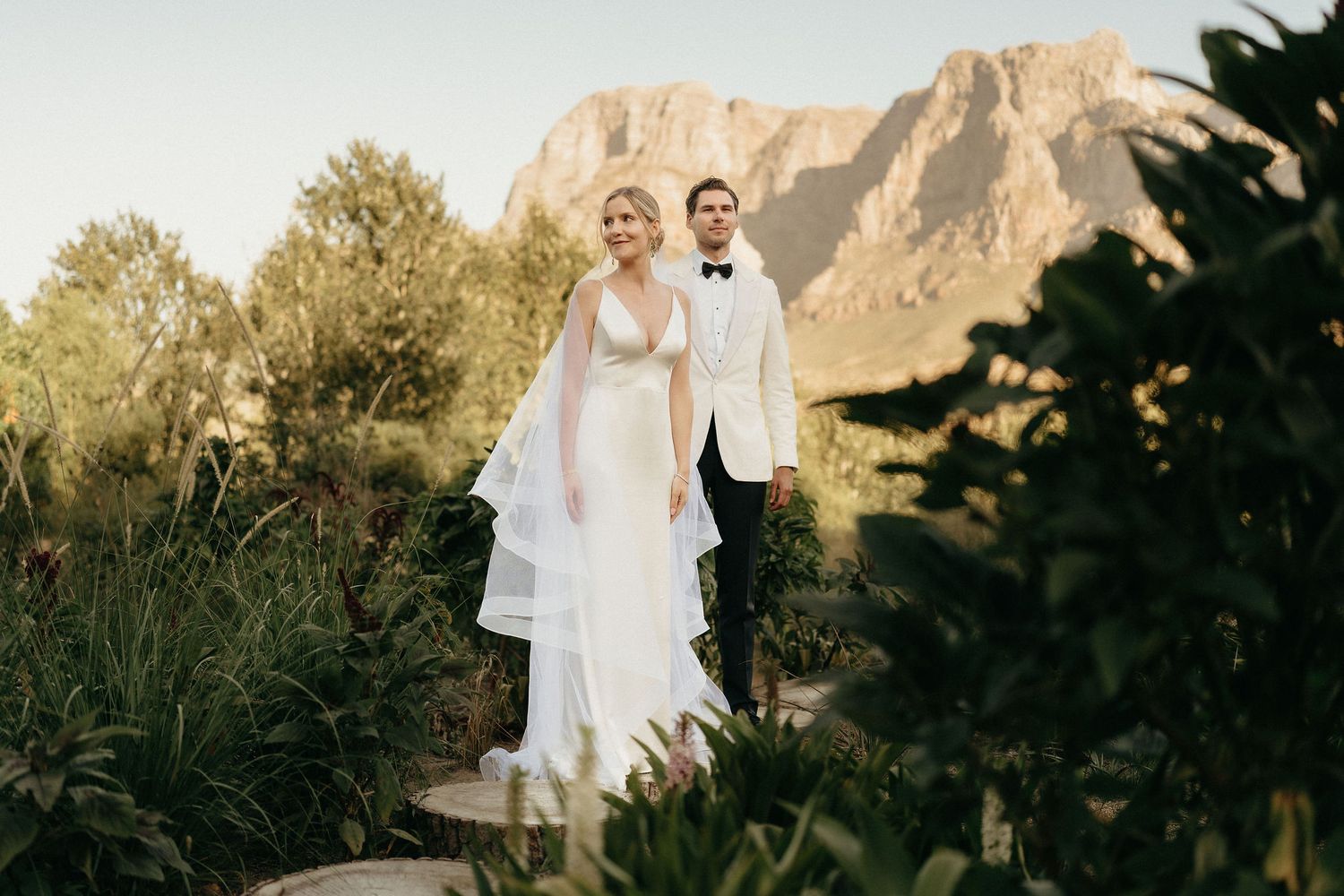 A couple in formal wedding attire poses against a scenic mountain backdrop during golden hour.