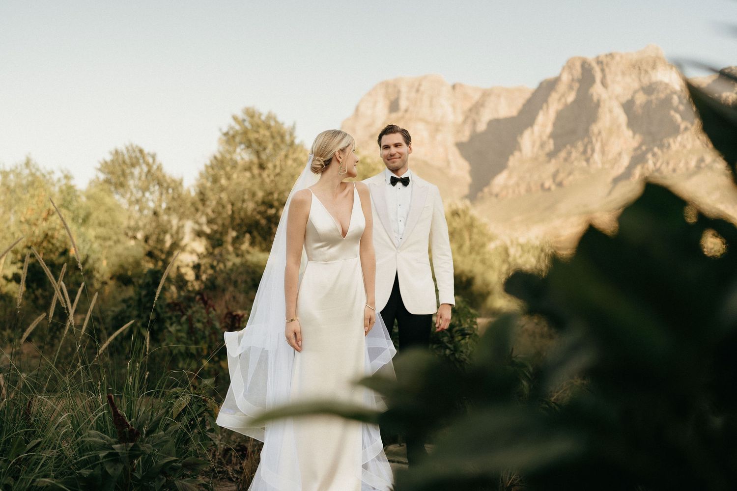 A couple in formal wedding attire embraces against a dramatic mountain backdrop at sunset.