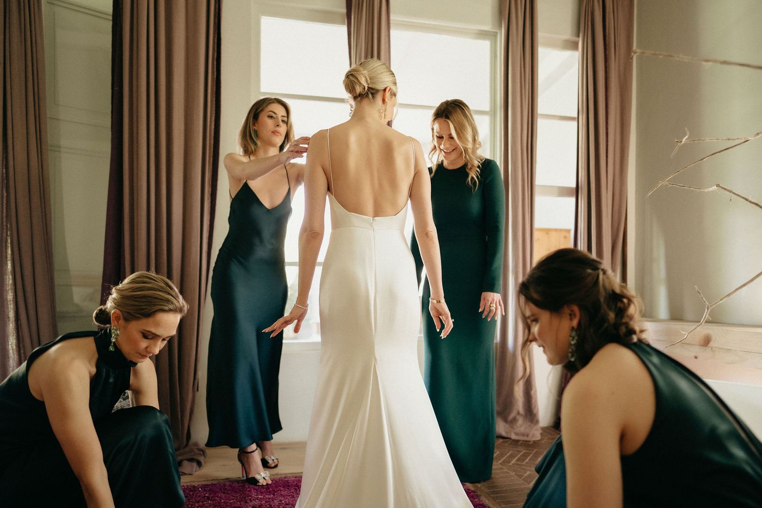 Bridesmaids in dark teal dresses help adjust a white wedding gown in a sunlit dressing room.
