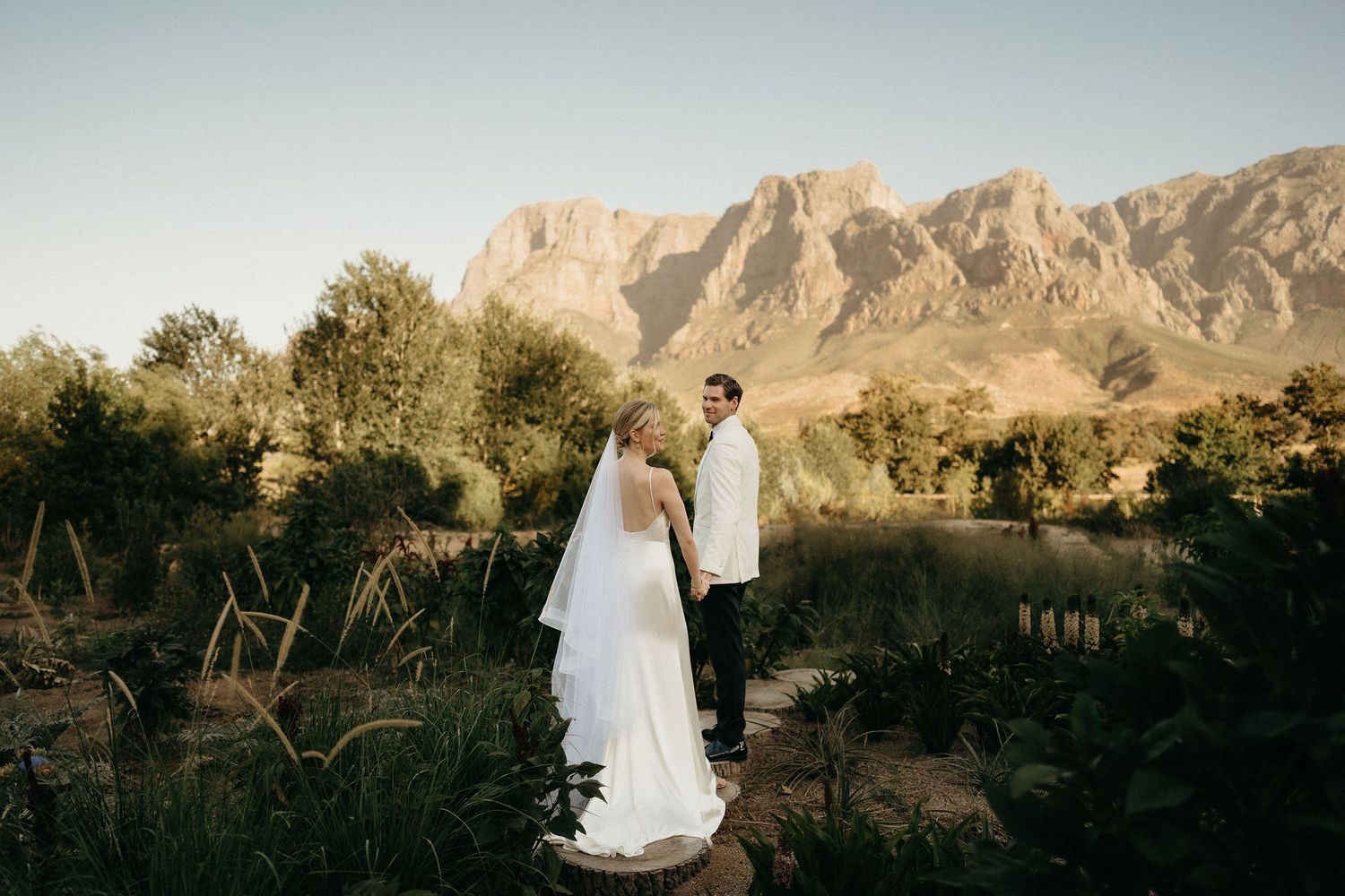 A couple in wedding attire stands together in a desert landscape with majestic mountains in the background at sunset.