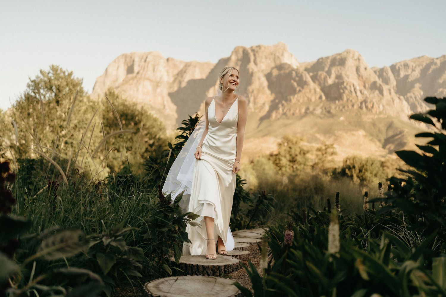 A bride in a flowing white dress stands gracefully against a backdrop of majestic desert mountains at sunset.