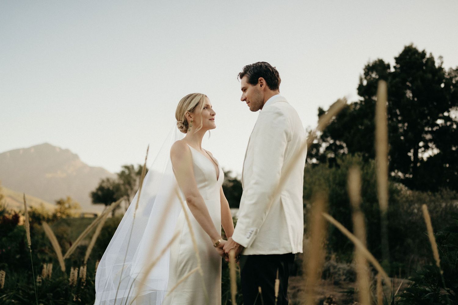 Couple in wedding attire embraces during golden hour photoshoot in a scenic mountain landscape.