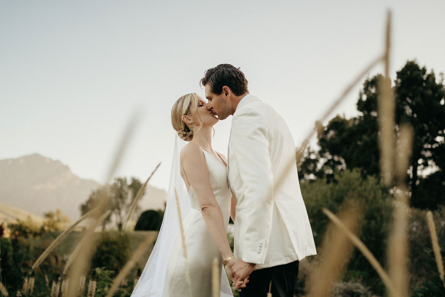 A couple in wedding attire shares a romantic moment in a golden-lit outdoor setting at sunset.