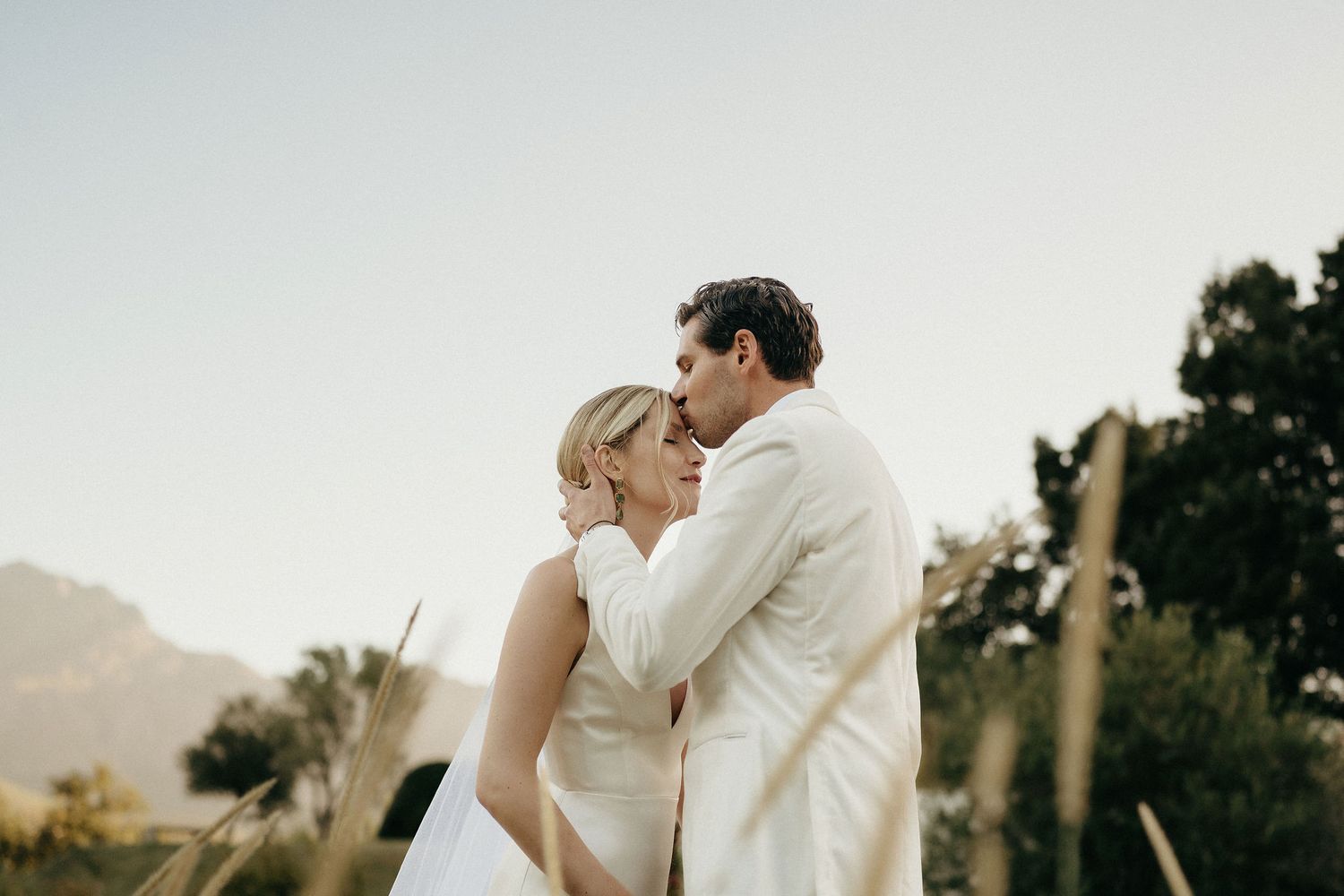 A couple in white wedding attire shares a romantic kiss during their outdoor mountain wedding ceremony.