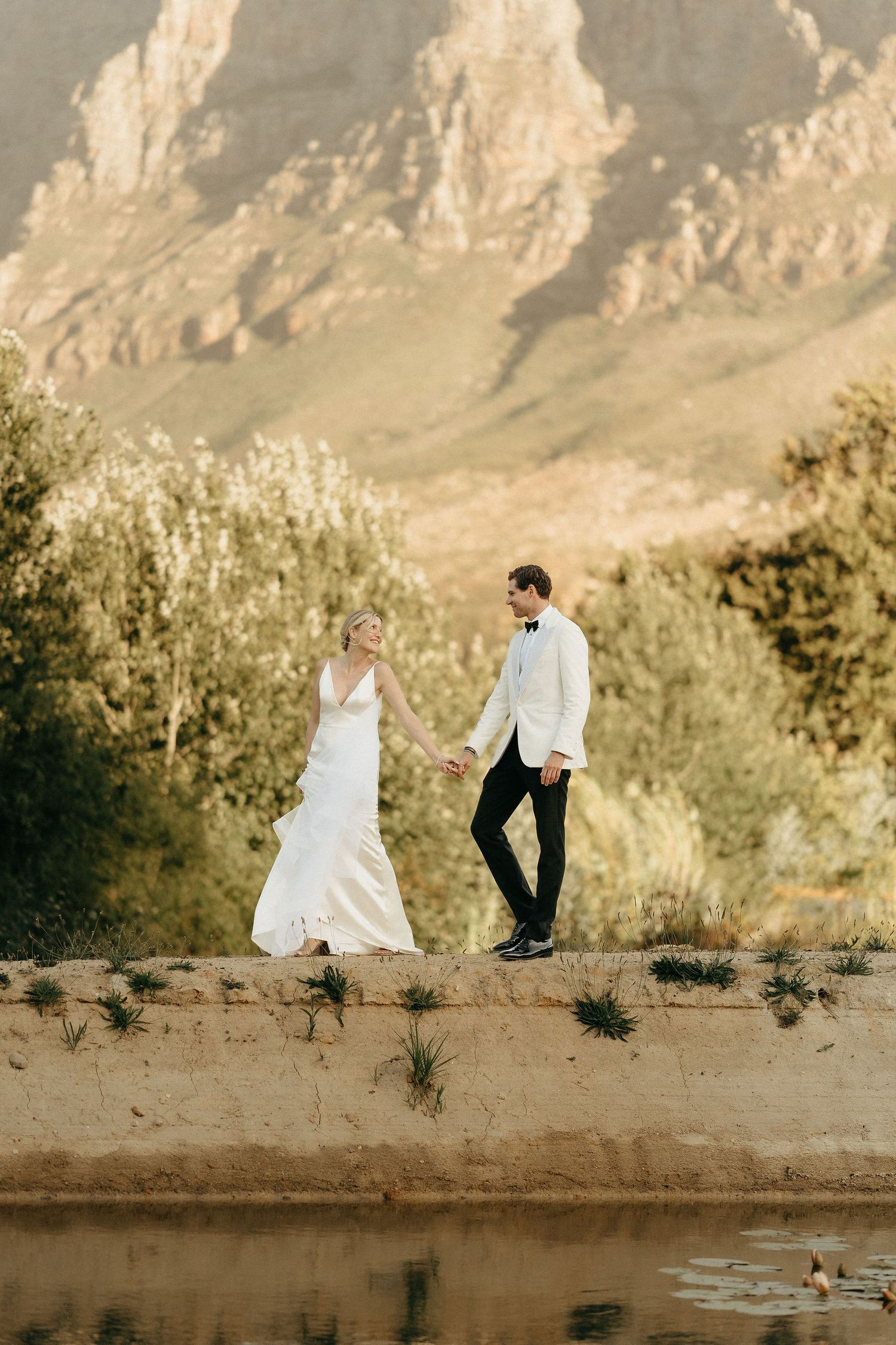 A couple in wedding attire walks along a scenic pond bank with dramatic mountain cliffs in the background.