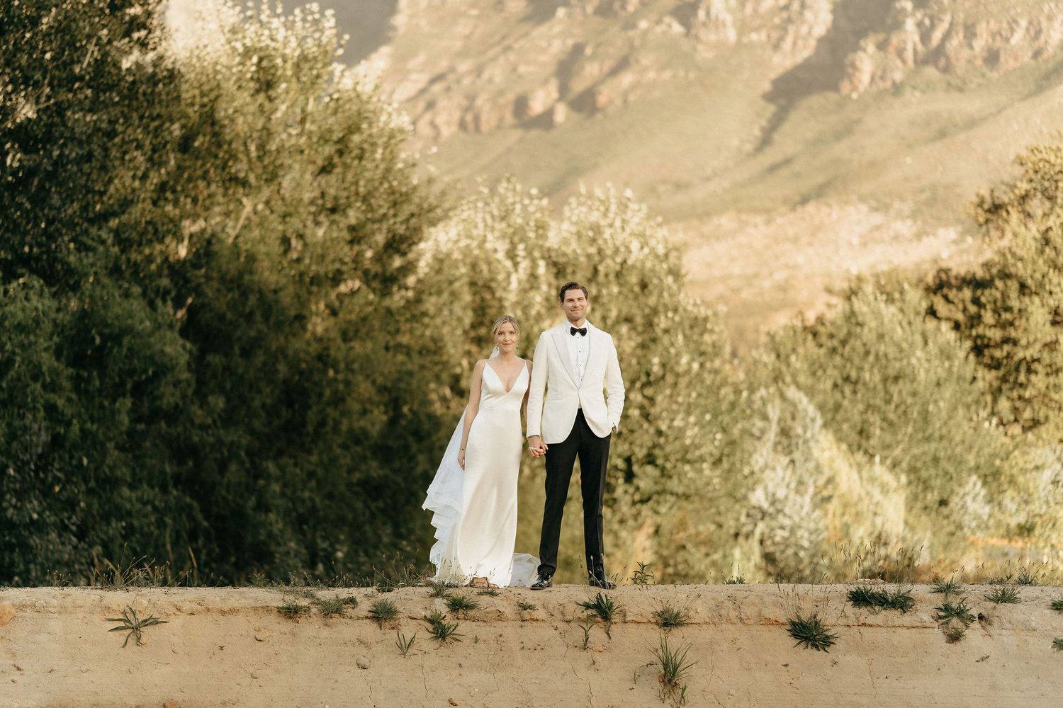 A couple in formal wedding attire stands together against a scenic desert mountain backdrop at sunset.