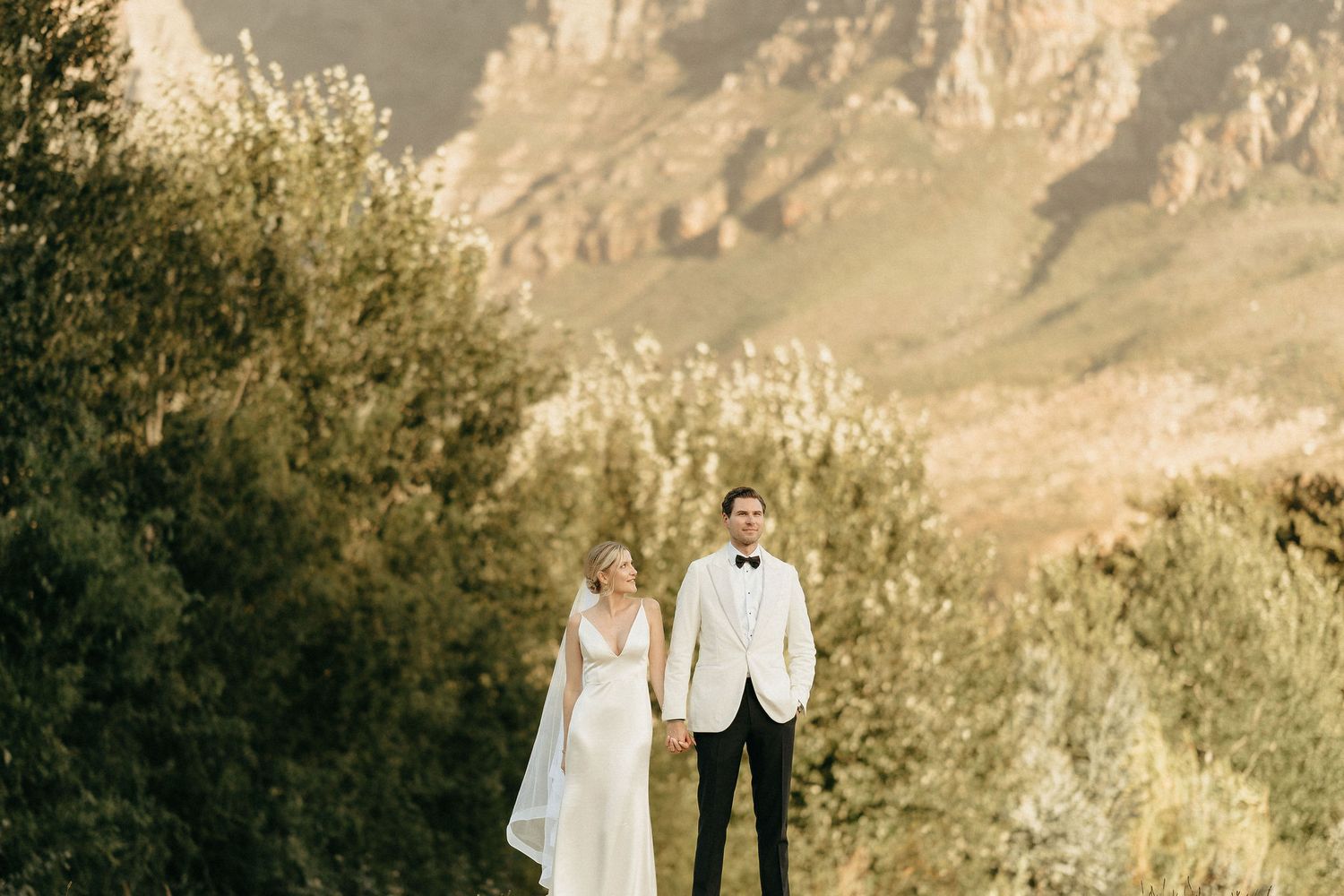 Couple in wedding attire stands together against dramatic mountain landscape in golden sunlight.