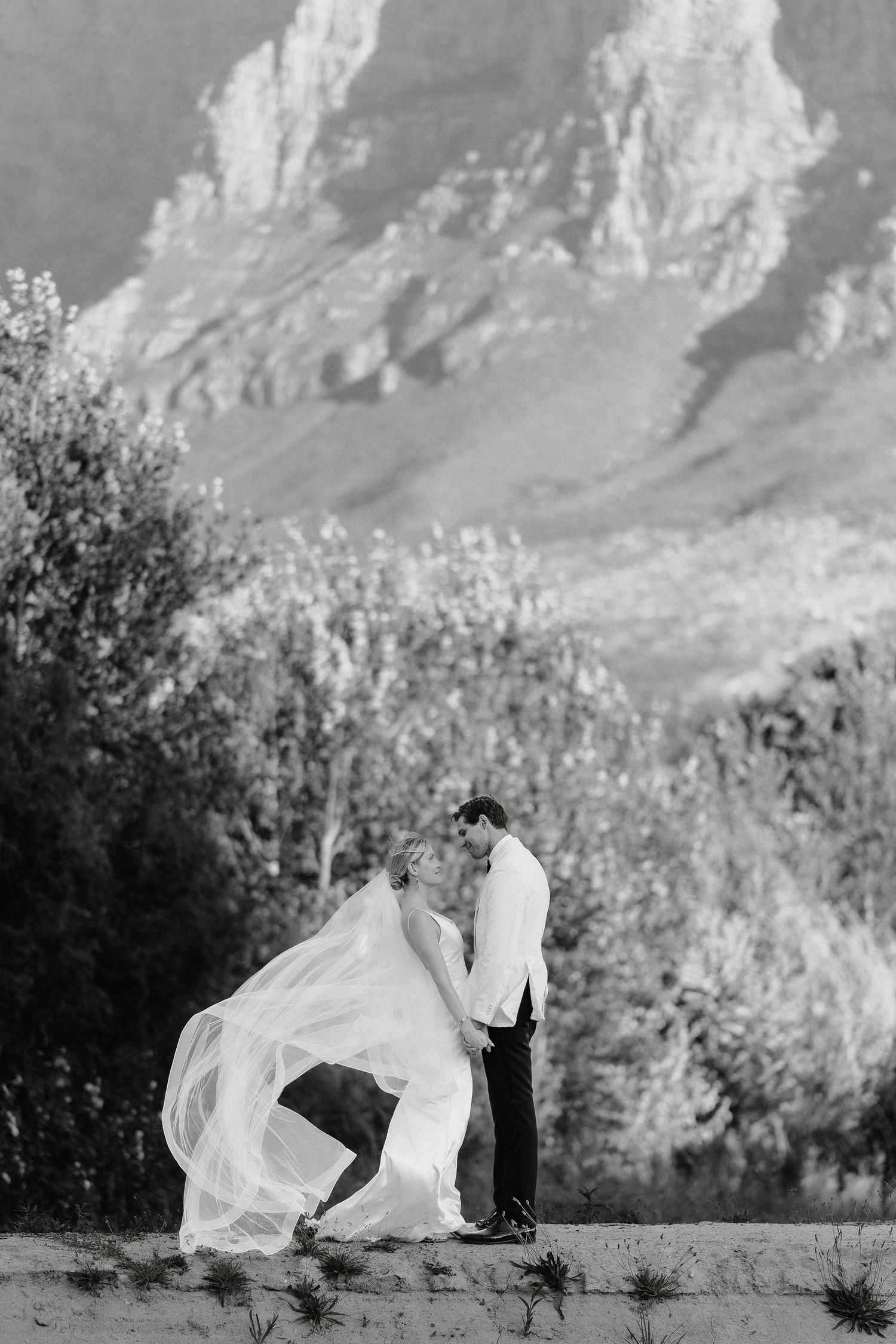 A romantic wedding couple poses against a dramatic mountain backdrop as the bride's veil flows in the wind.