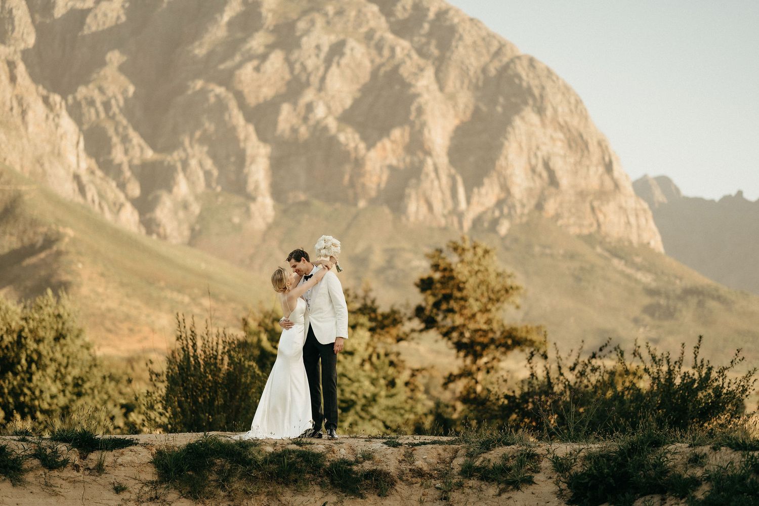 A couple in wedding attire embraces against a dramatic mountain landscape during golden hour.