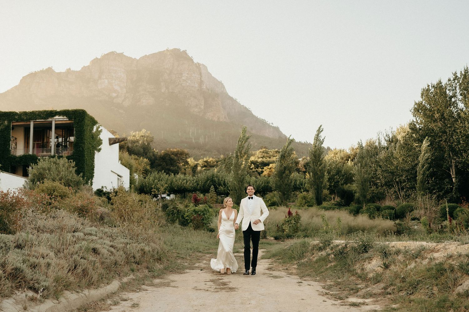 A couple walks down a dirt path with a mountain and rustic building in the background during golden hour.