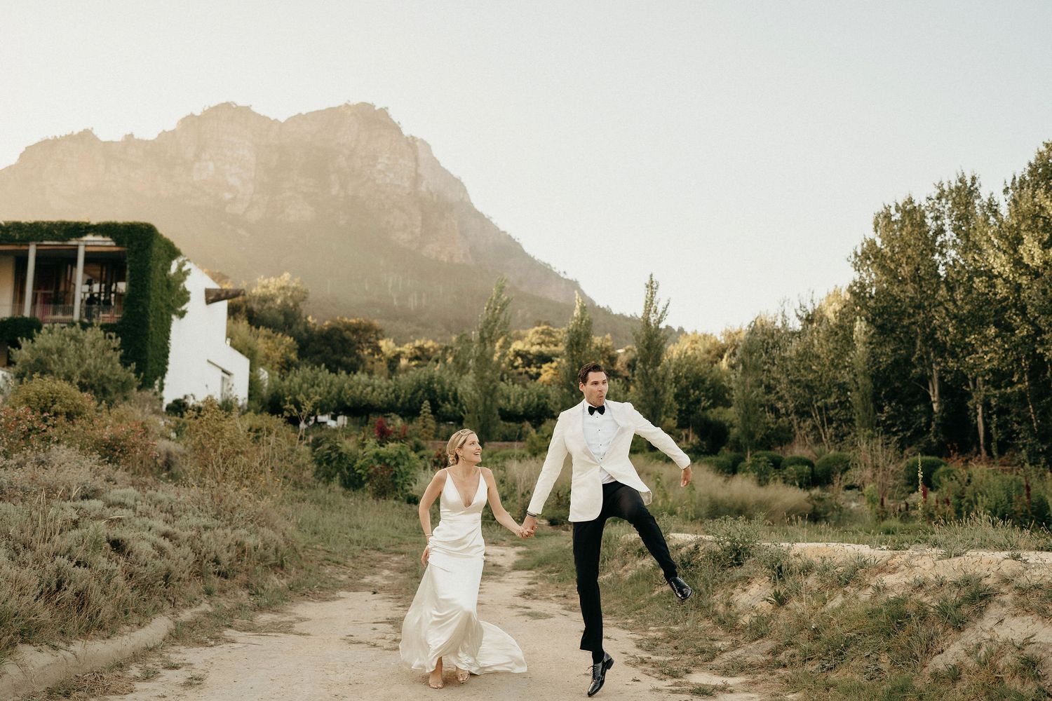 A couple in wedding attire walks through a scenic mountain landscape at sunset.