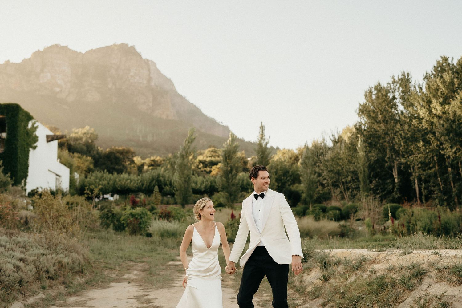 A couple walks hand in hand through a desert landscape with mountains and saguaro cacti in the background at sunset.