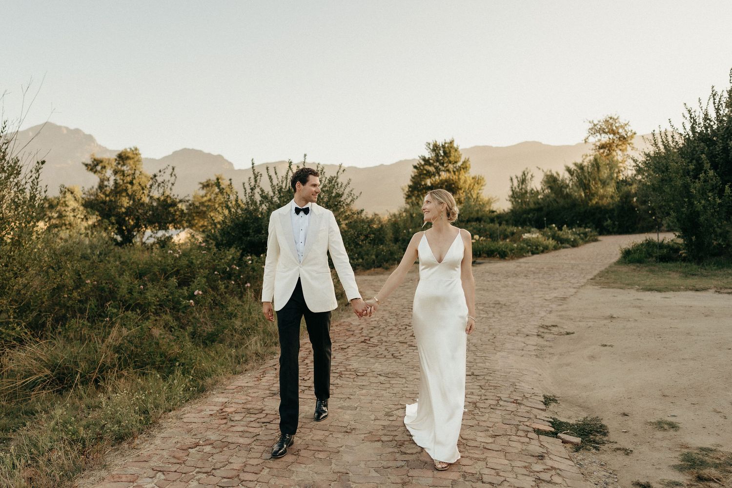 A couple walks along a desert path during golden hour with mountains in the background.