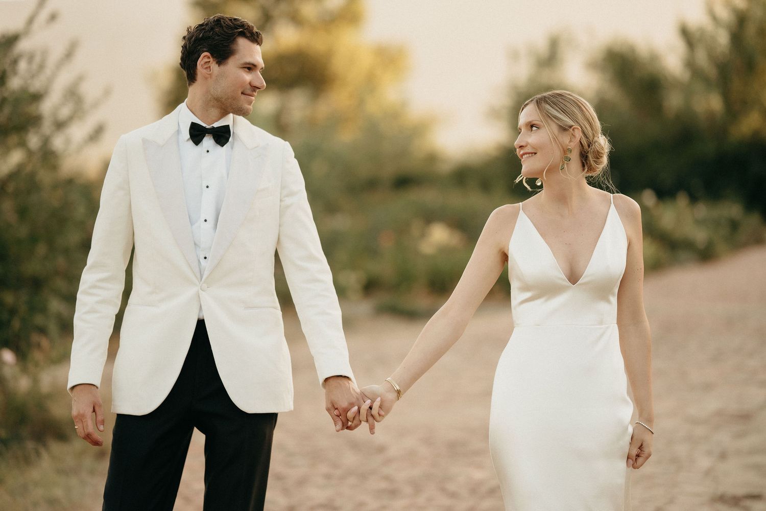 A couple in wedding attire holds hands while walking through a desert landscape at sunset.