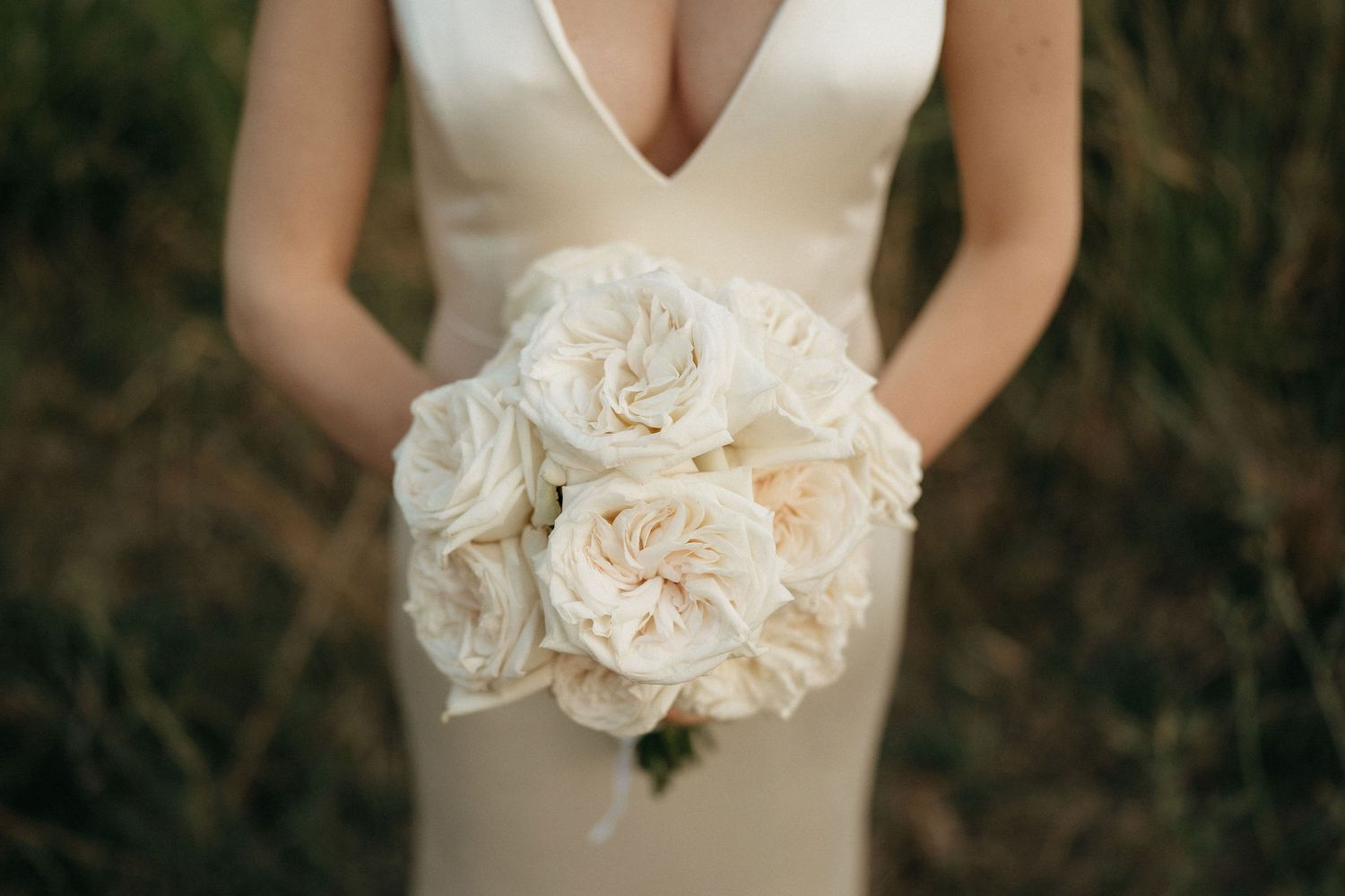 A bride in a sleek white dress holds a romantic bouquet of cream and blush roses against a natural outdoor background.