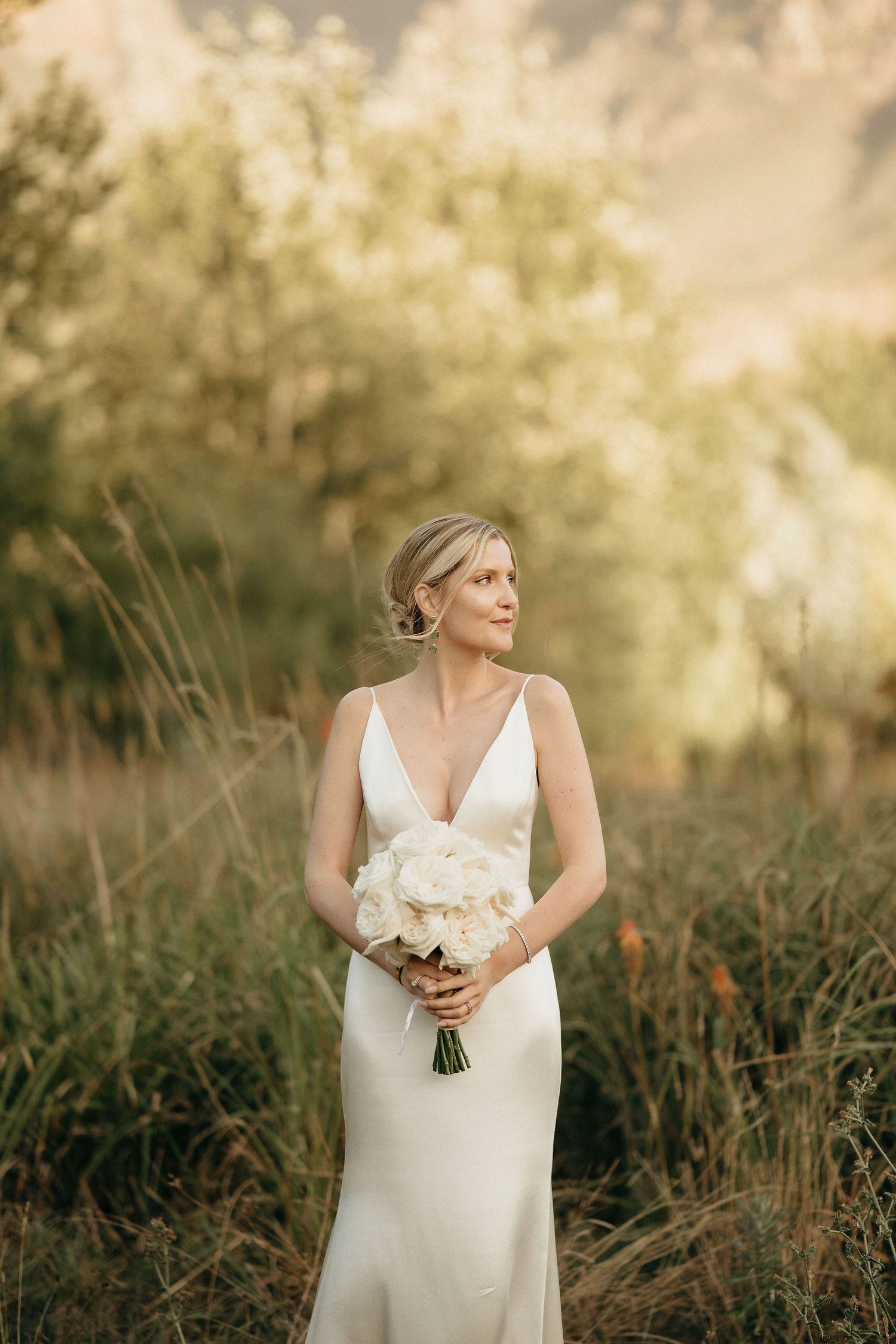 A bride in an elegant white v-neck gown holds a white bouquet while standing in tall grass during golden hour.