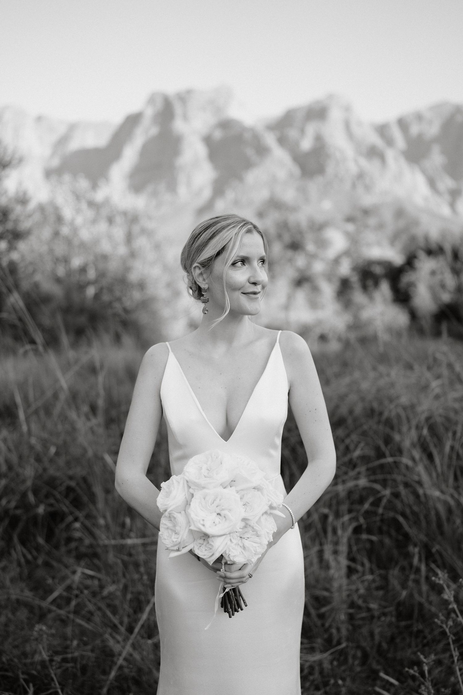 Black and white portrait of a bride in a sleek v-neck wedding dress holding a white bouquet with mountains in background.
