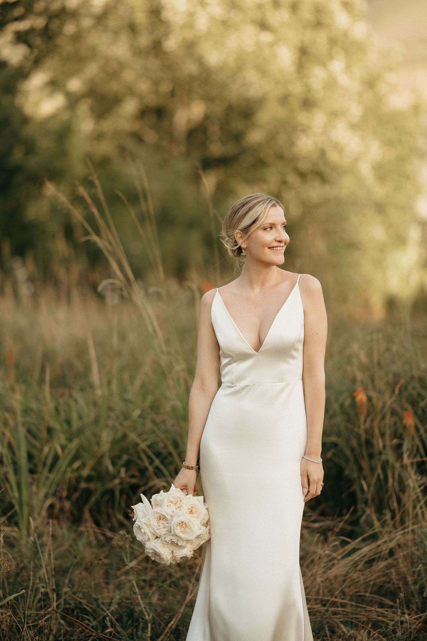 A bride in a sleek white V-neck wedding dress poses gracefully in a natural outdoor setting with tall grass and soft lighting.
