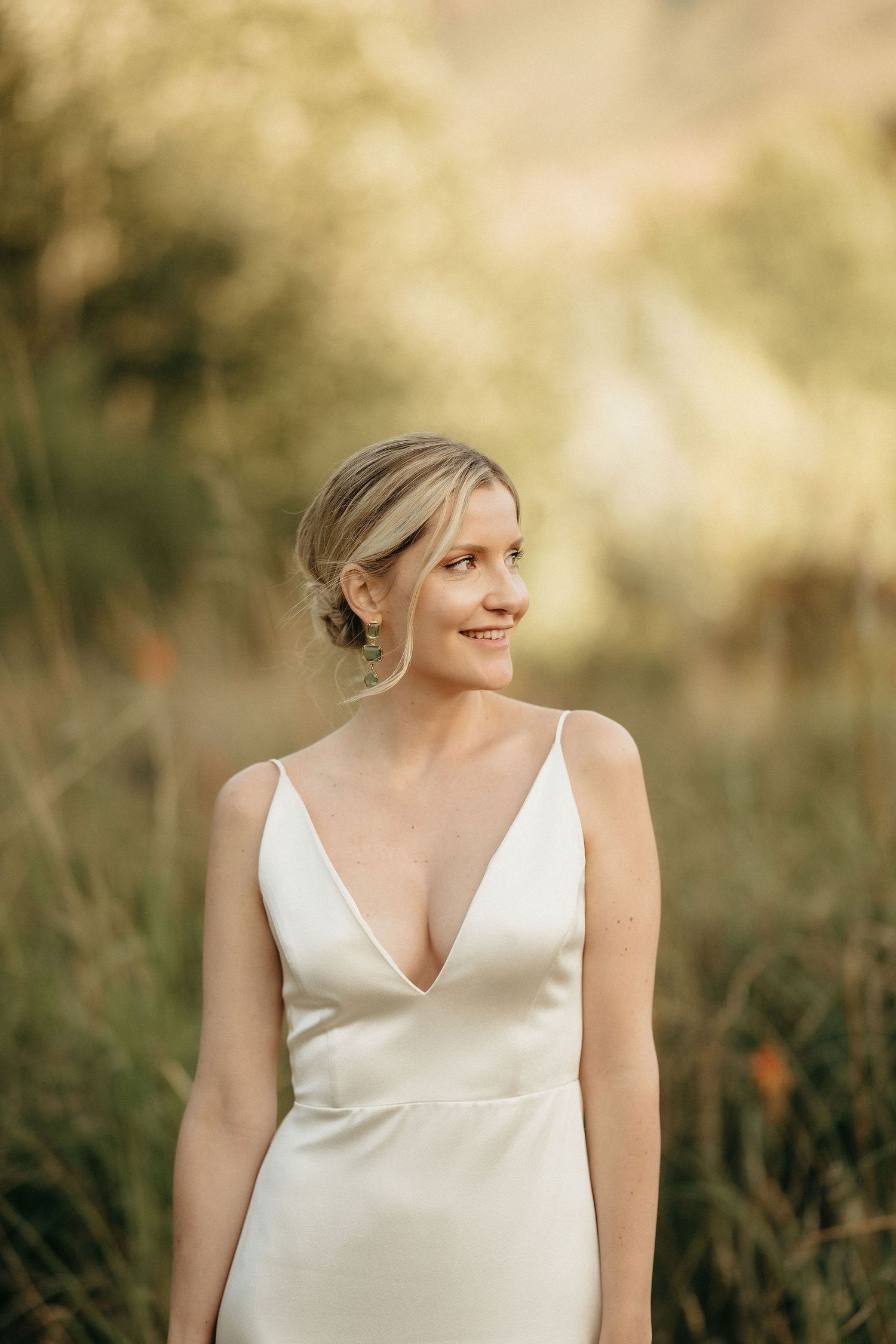 A person in a white sleeveless dress poses outdoors in a natural setting with soft bokeh lighting.
