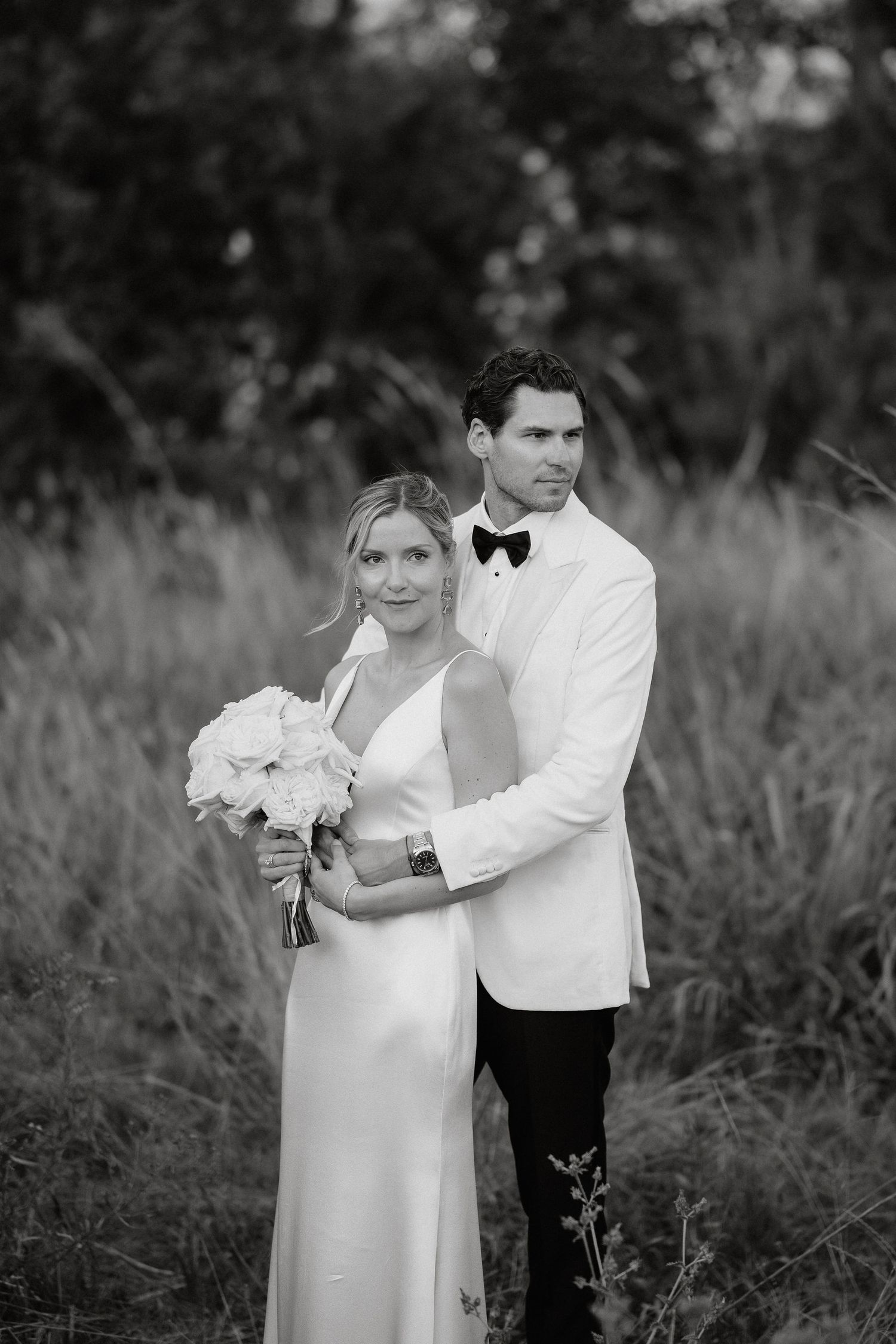 A black and white wedding photo series shows a couple embracing in a natural outdoor setting with tall grass.