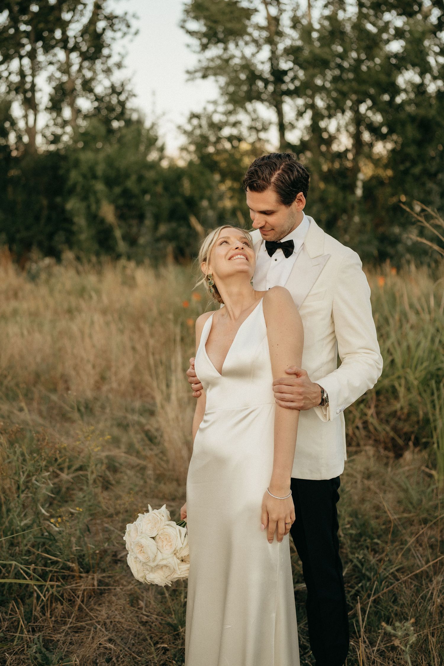 A romantic wedding couple embraces in a natural outdoor setting with golden light filtering through pine trees.