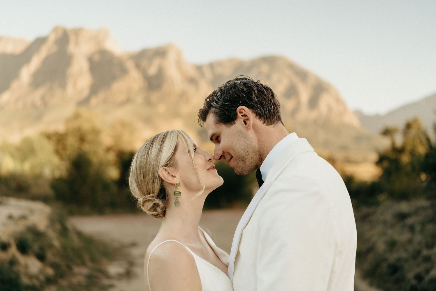 A couple in white clothing shares a romantic moment against a backdrop of desert mountains at sunset.