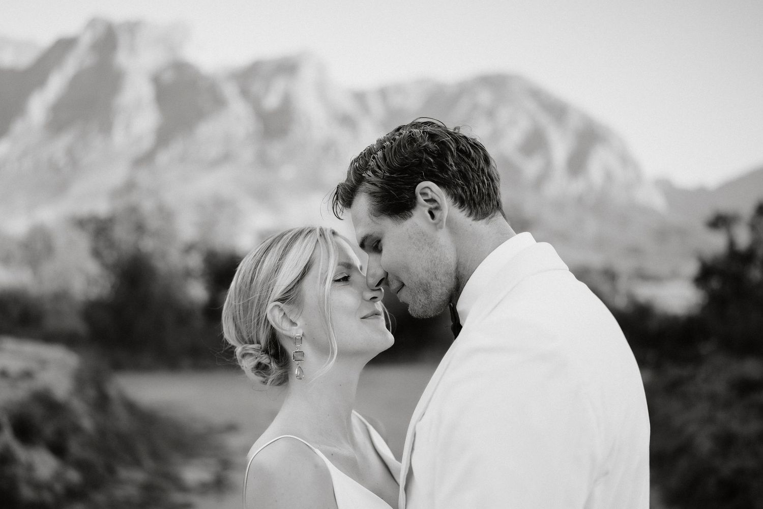 A romantic couple shares a tender moment against a scenic mountain backdrop in black and white.