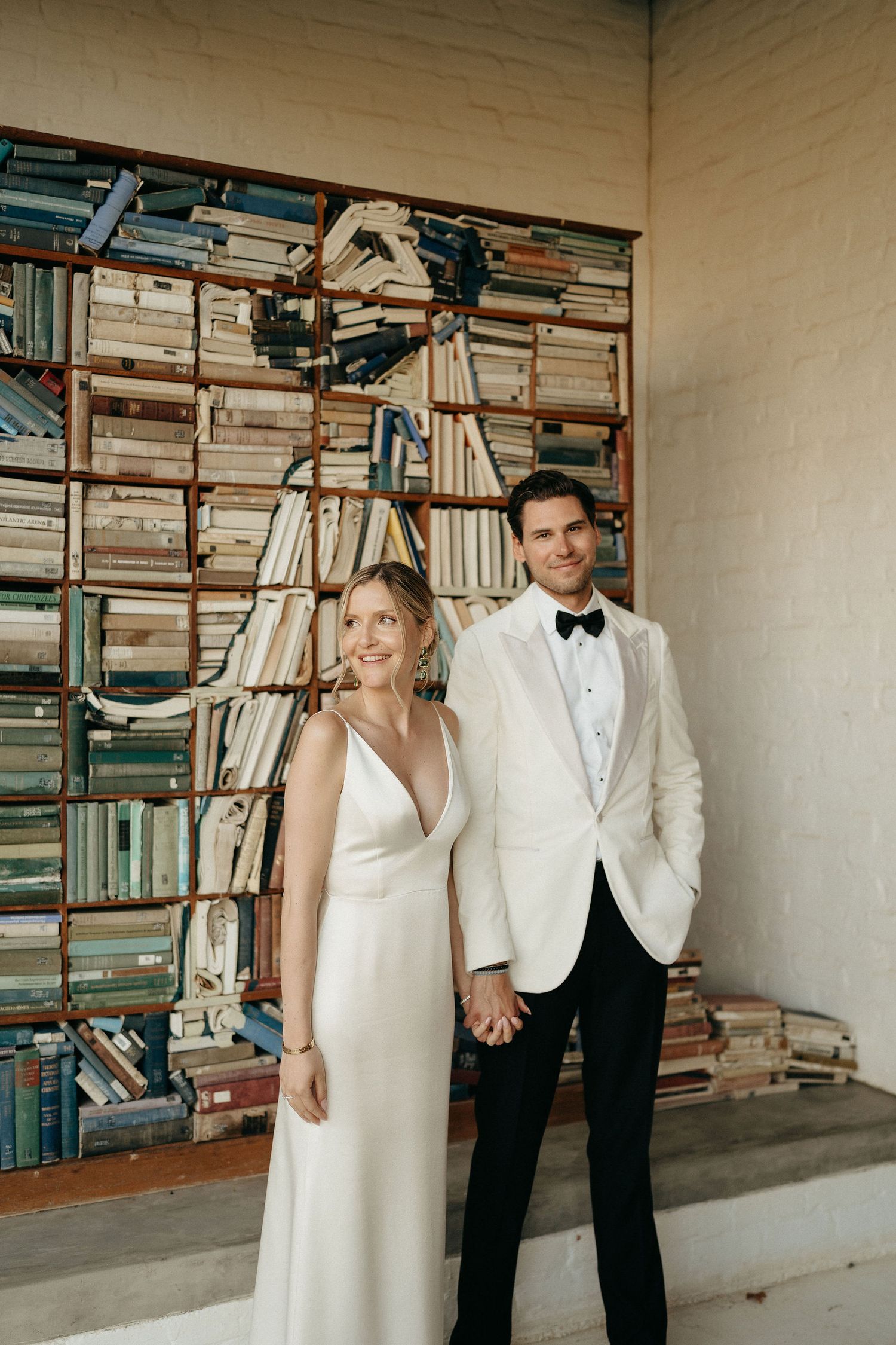 A couple in formal wedding attire stands in front of a wall of stacked books and bookshelves.