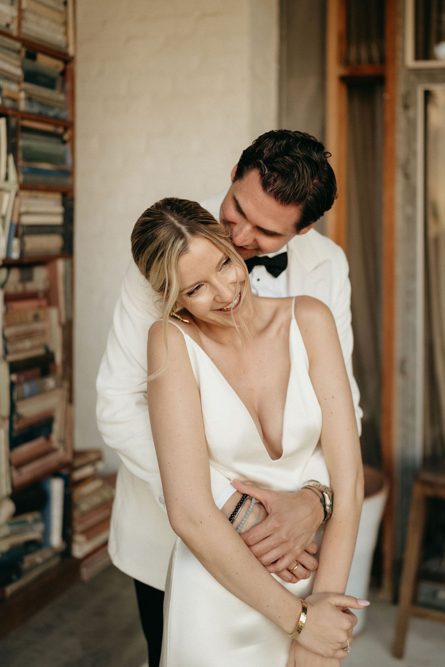 Romantic wedding couple embraces in intimate moment wearing white formal attire against rustic backdrop.