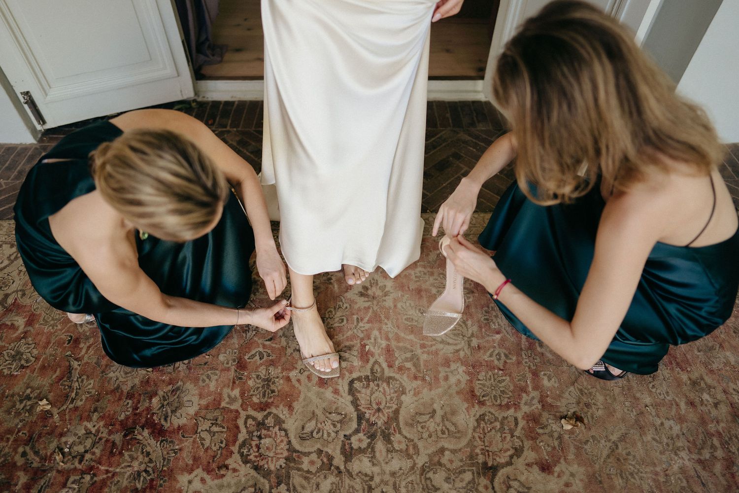 Two people in teal dresses help adjust a white dress and shoes on an ornate red carpet.